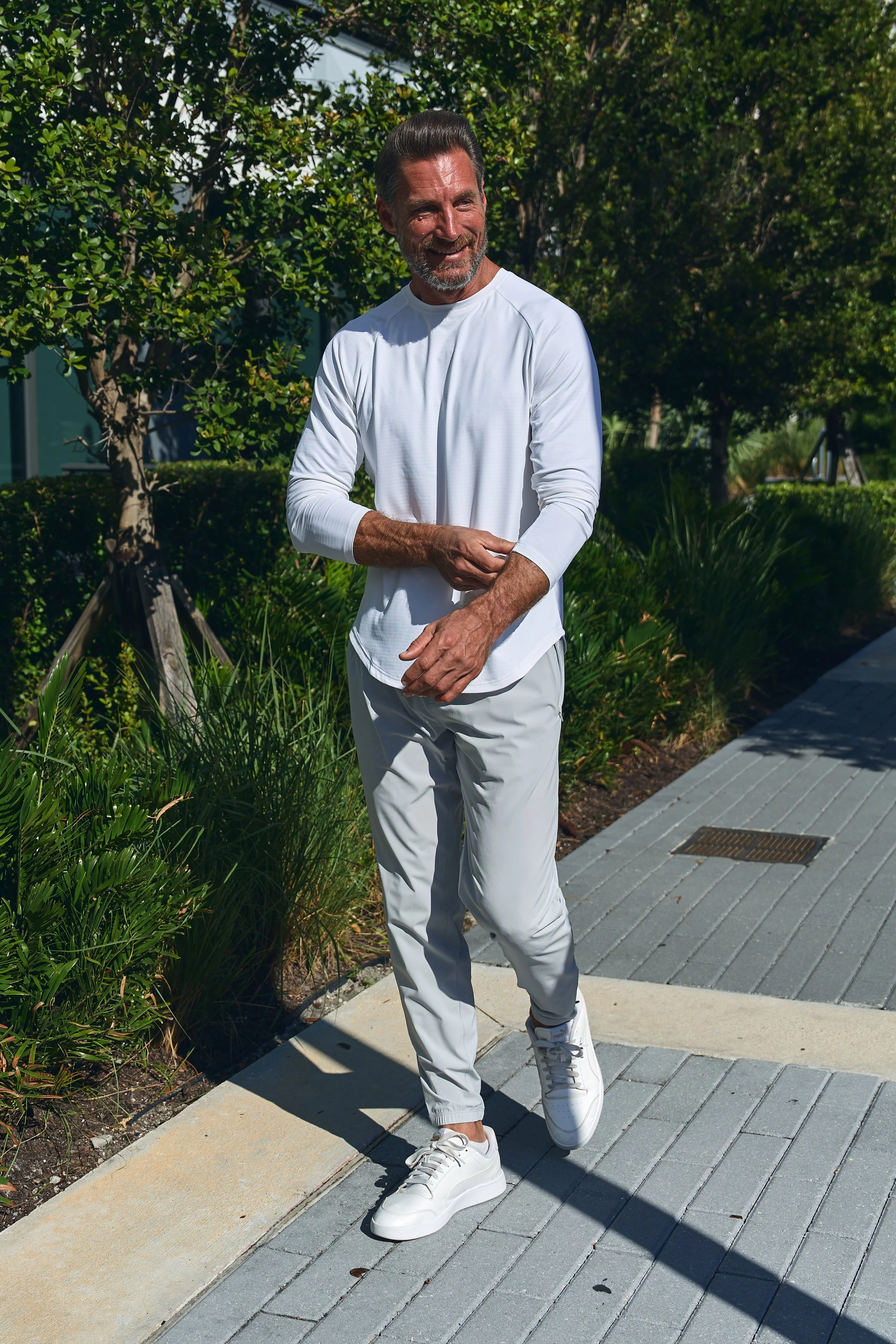 A man walking on a paved sidewalk outdoors, wearing a white long-sleeve shirt, light gray pants, and white sneakers, smiling and enjoying sunny weather with green trees and bushes in the background.