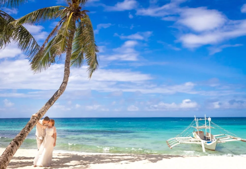 Tropical beach with a bride and groom posing underneath a palm tree.