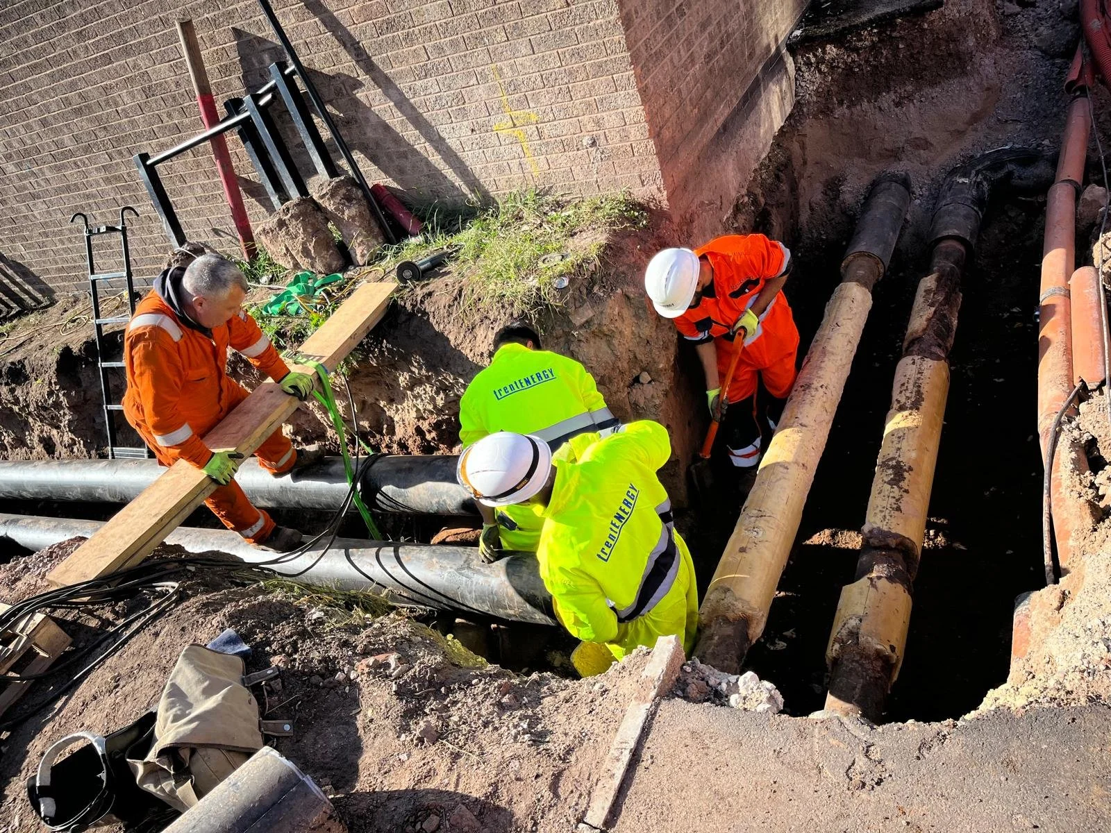 Workers in safety gear repairing or installing pipes in a trench alongside a brick wall.