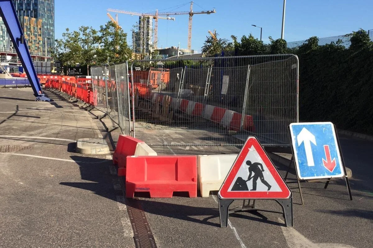 Road construction site with barriers, warning signs, and fencing, with cranes and buildings in the background.