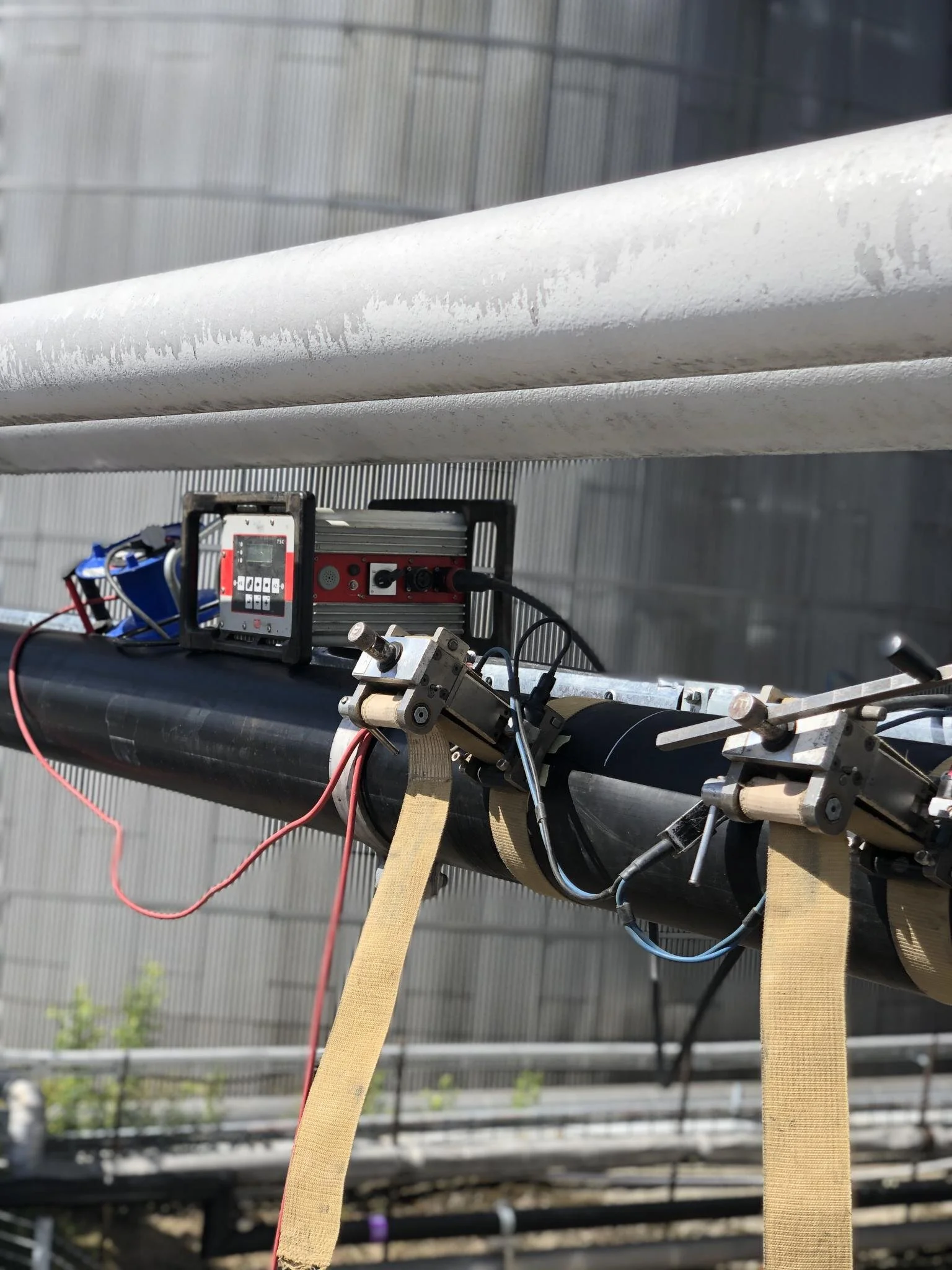 Close-up of a black pipeline with attached sensors and electronic equipment, secured with beige straps, outdoors on a sunny day.