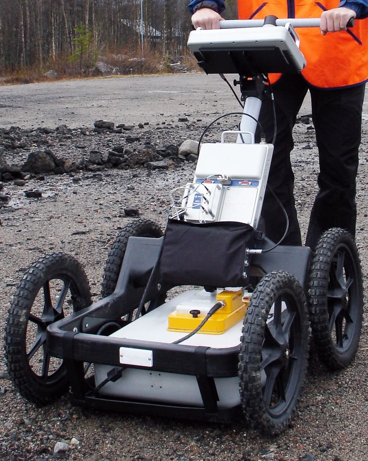 A person in an orange jacket and black pants operating a robotic vehicle on a gravel surface in a forested area.
