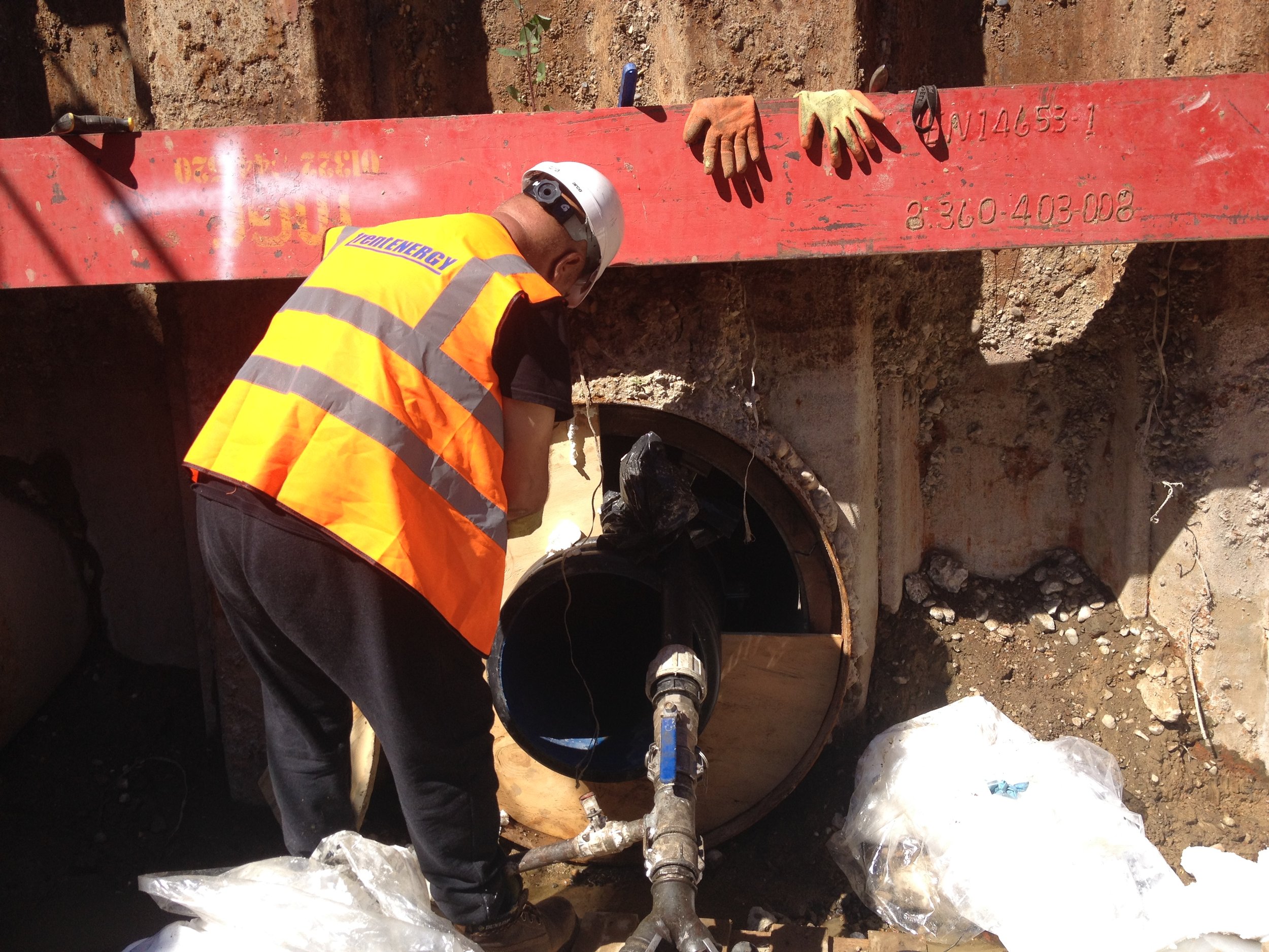Construction worker wearing a yellow safety vest and a white helmet working on a large underground pipe at a construction site.