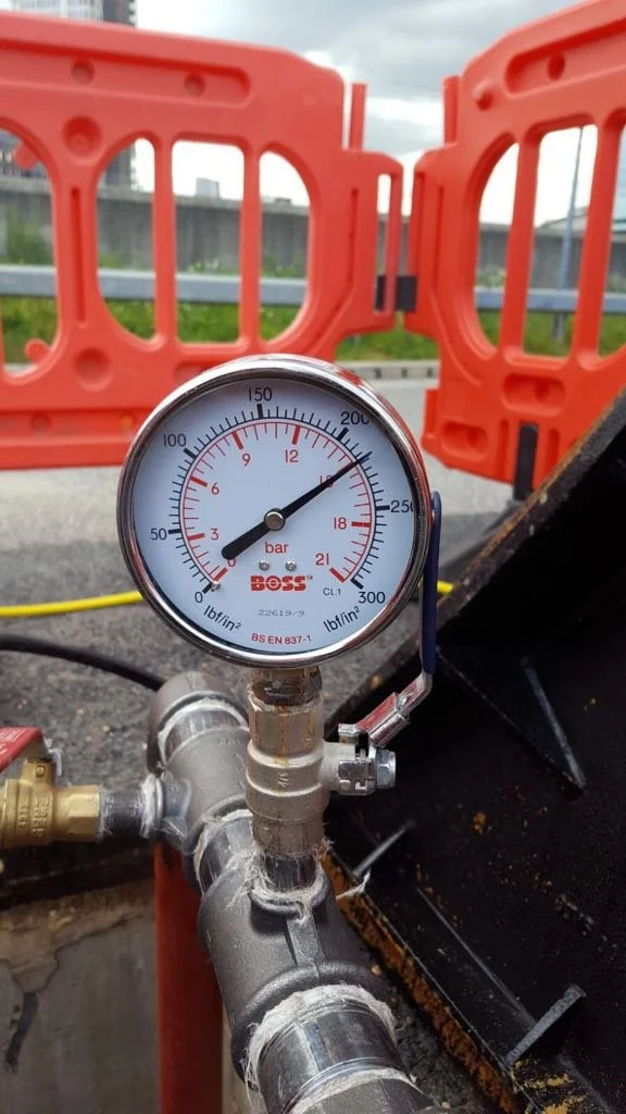 A pressure gauge attached to piping on a construction site, with orange barriers and a cloudy sky in the background.