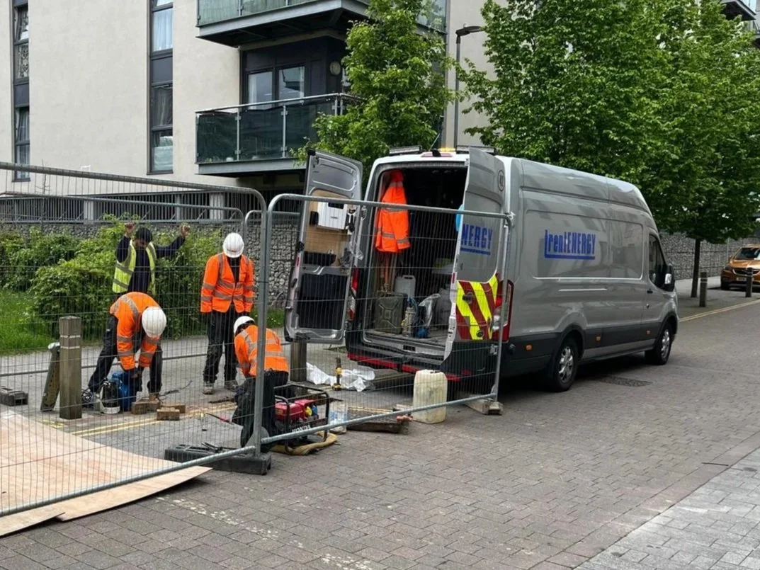 Construction workers wearing orange safety vests and white helmets working on a street near a fenced construction site with a grey utility van parked nearby.
