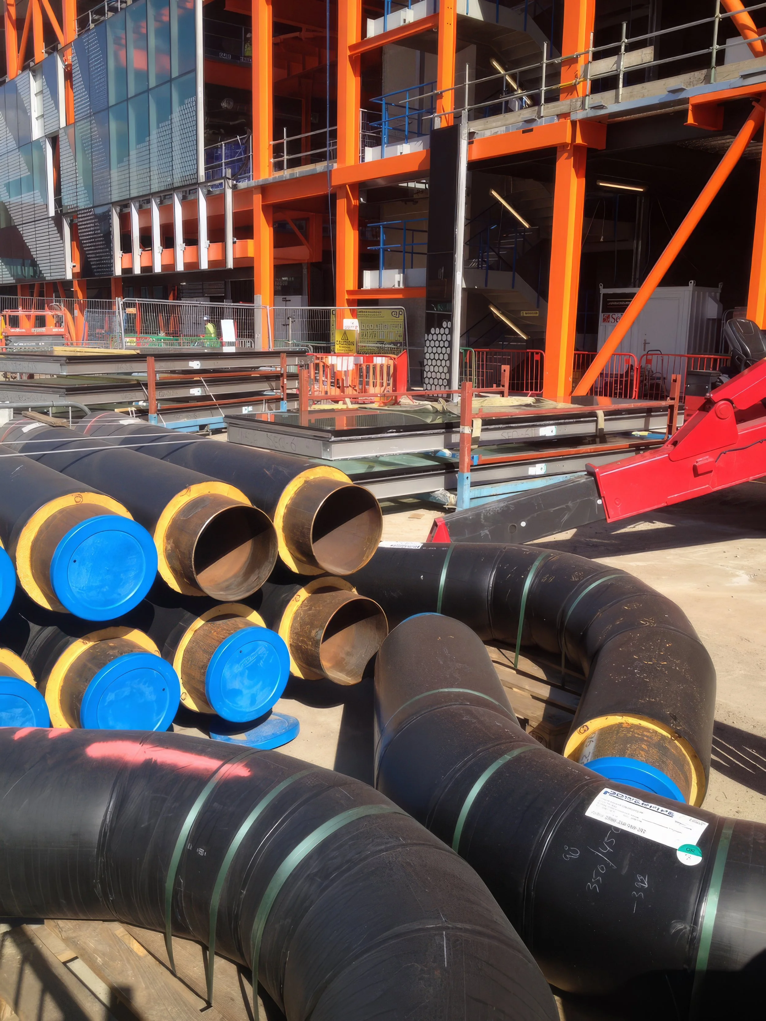 Construction site with large black pipes with blue and yellow caps, and orange structural elements of a building under construction.