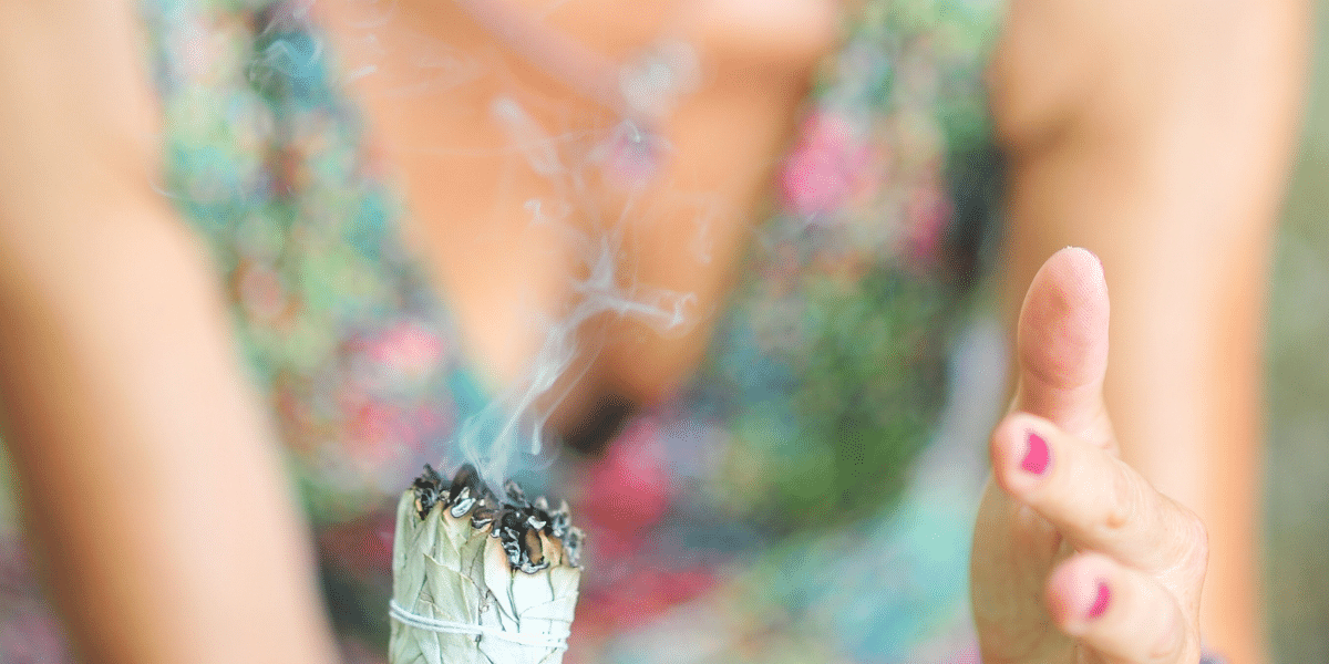 Une personne fait brûler un bâton d'encens, avec de la fumée qui s'élève, devant un arrière-plan flou de vêtements colorés.