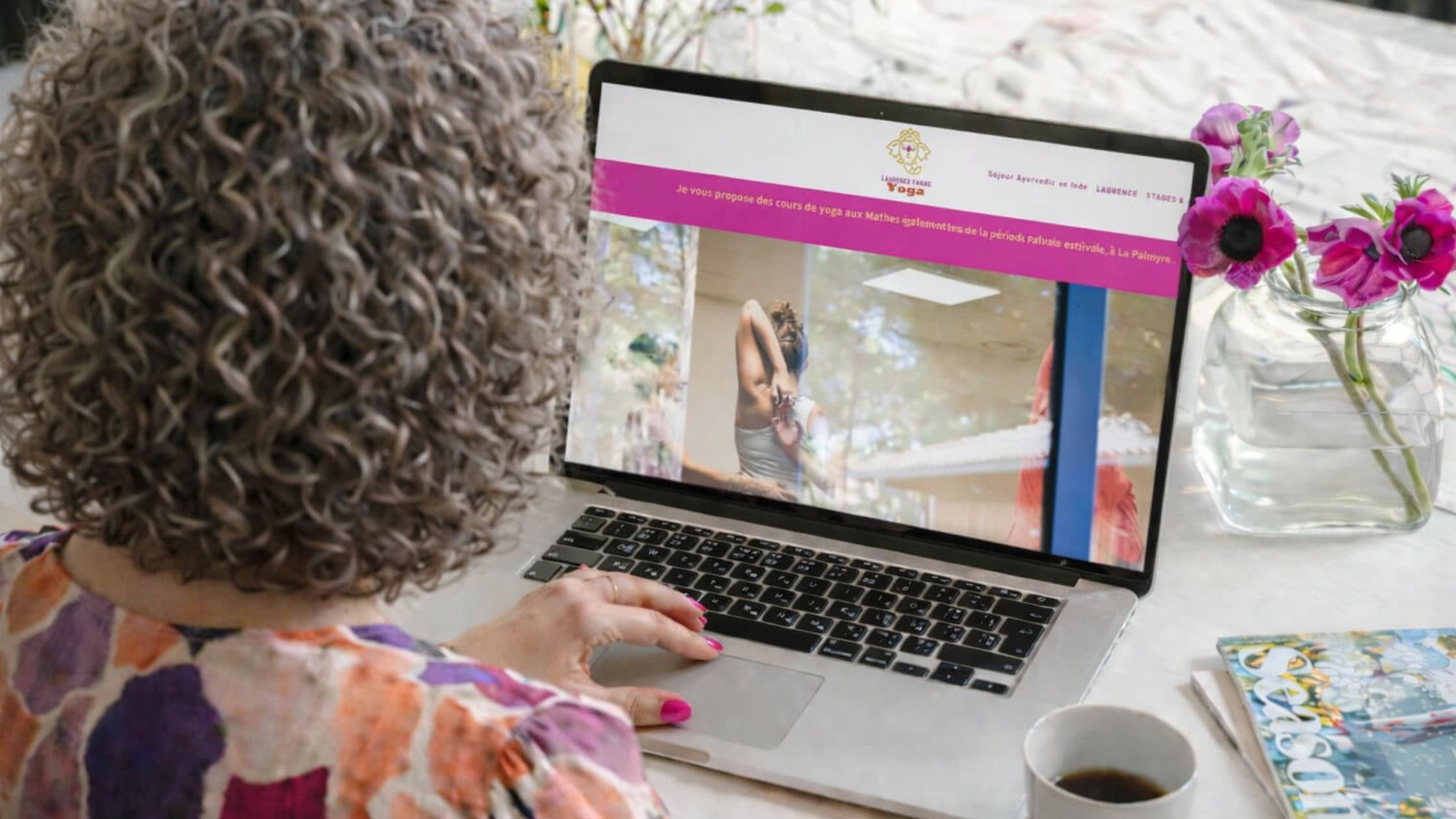 Une femme avec des cheveux bouclés assise devant un ordinateur portable, regardant un écran qui affiche une page de site web ou de publicité avec une image d'une femme dans une pose de yoga; une vase avec des fleurs roses est visible à droite, et une tasse de café est en bas à droite.