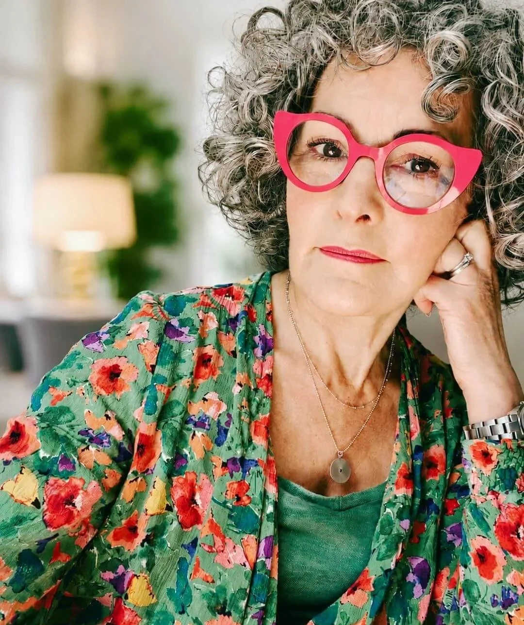 Femme aux cheveux gris bouclés portant de grandes lunettes roses, un chemisier à motifs floraux colorés, et un collier en chaîne avec un pendentif, posant de façon pensive dans un intérieur lumineux.