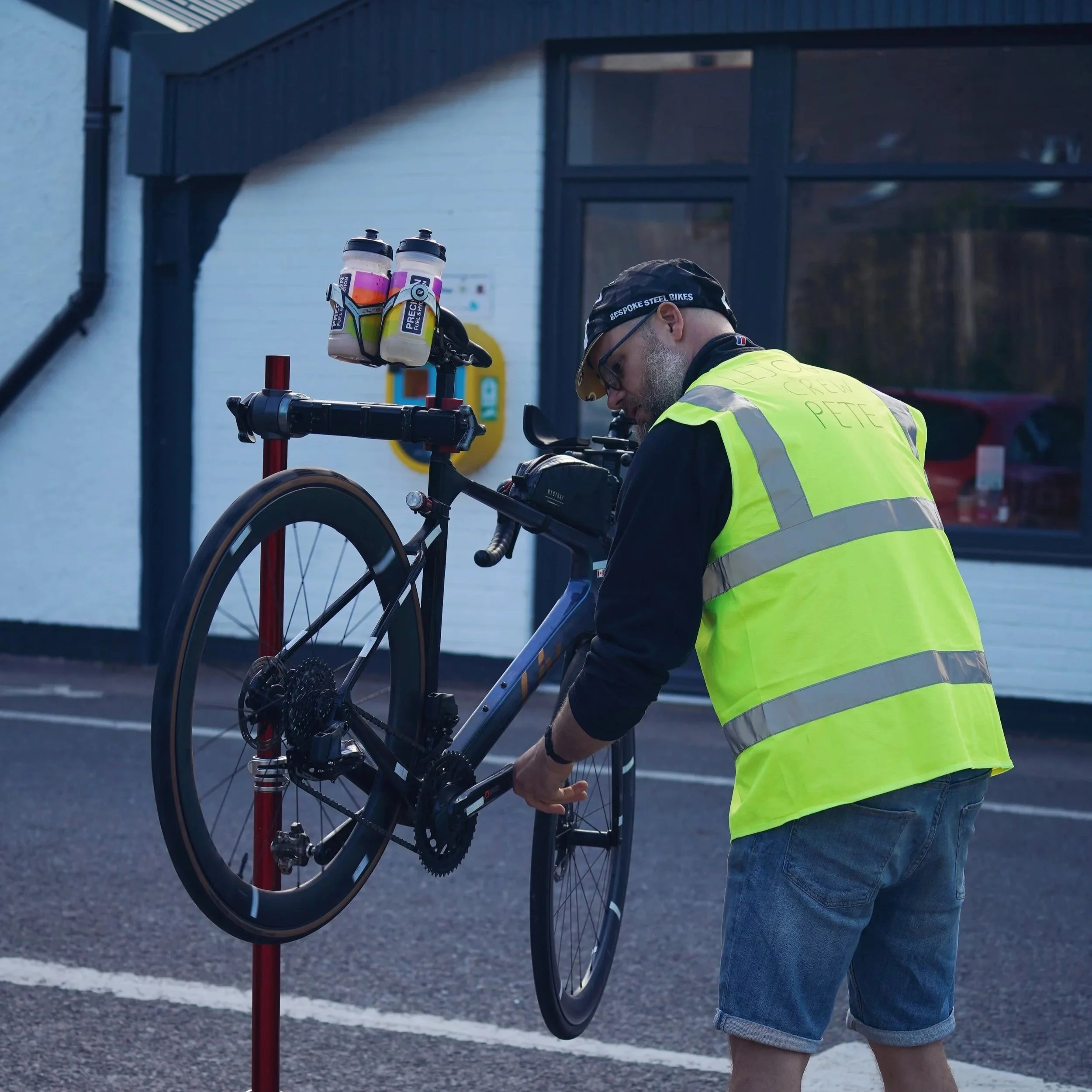 A man in a yellow safety vest adjusting a black bicycle on a bike repair stand outside a building with white walls and large windows.