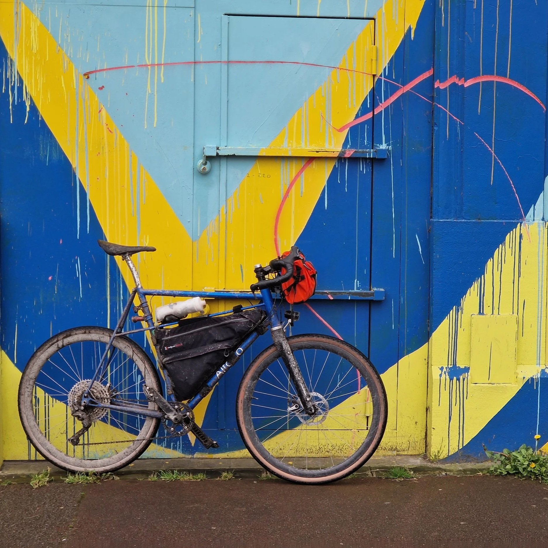 A mountain bike leaning against a colorful blue and yellow geometric mural on a wall. The bike has a red handlebar bag and a black frame bag, with a white water bottle attached. The ground is wet and the mural features paint drips.