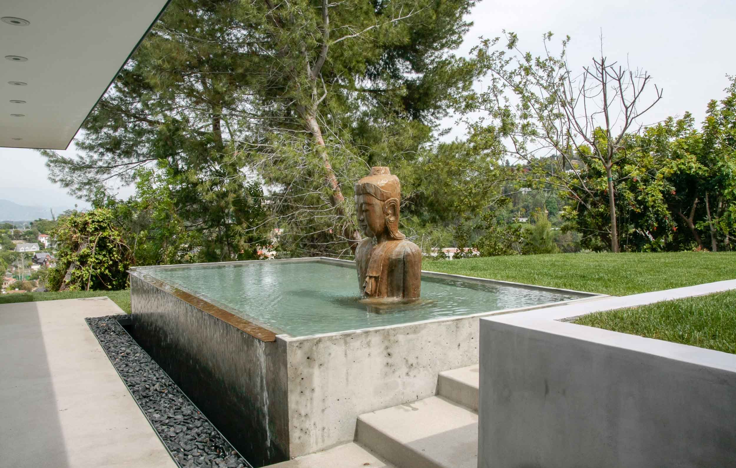 BLUM RESIDENCE LANDSCAPE A typical grass rear yard was transformed into a place for both entertaining and meditation. These conflicting functions were resolved by creating two areas separated by a change in level. A concrete water pond was placed at this transition. The pond edge brings the soothing sound of falling water into the backyard. Steps alongside the pond lead to a stone path taking one to the meditation area at the rear of the yard. A white ceiling adjacent to the house wraps around and defines the entertaining area. A built-in grill and long seating counter define one end of this area. Studio City, CA