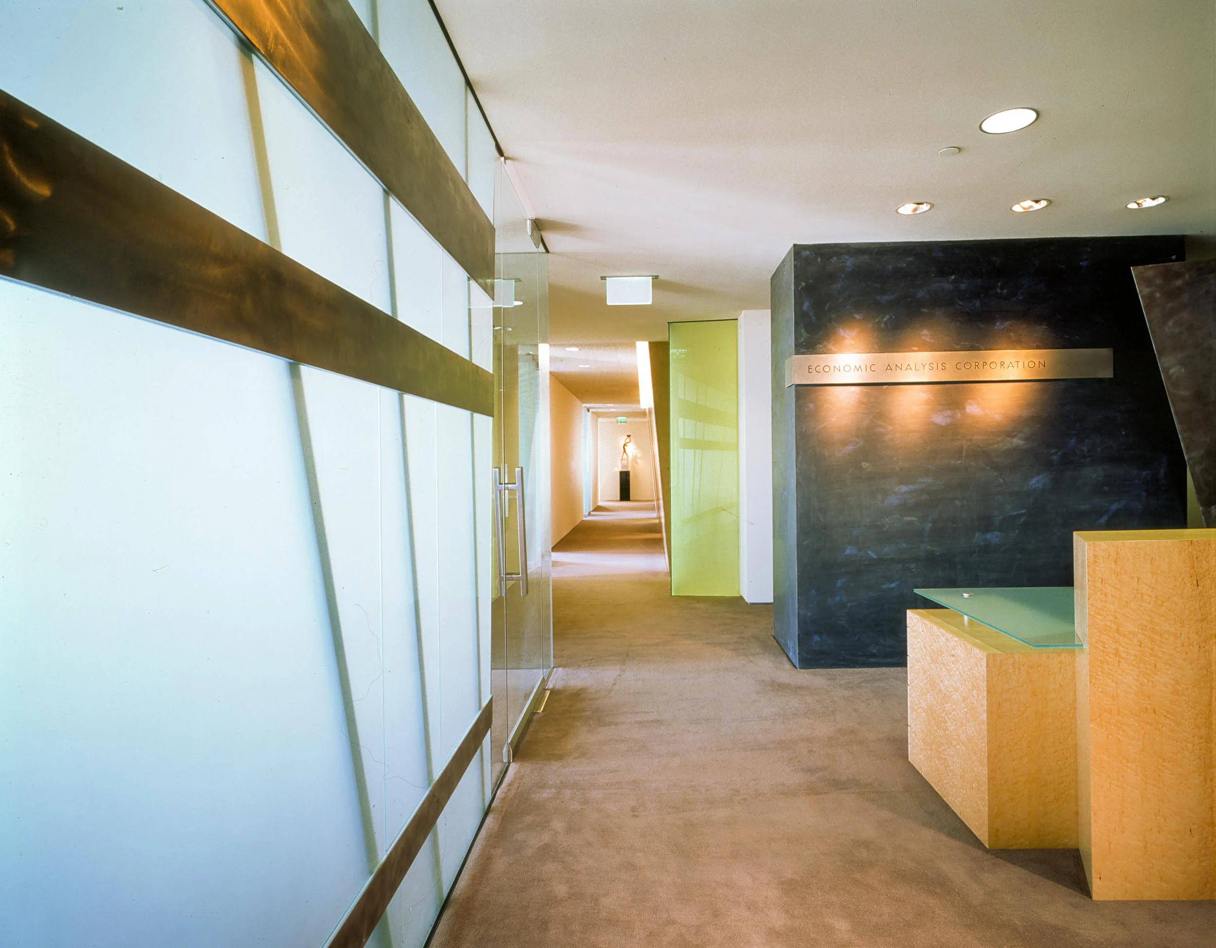 ECONOMIC ANALYSIS CORPORATION Opposing and contrasting forms, materials and color are held in harmonious balance in such a way as to create an overall experience of resolution, balance and repose contained within an envelope of serenity. Separate but related elements present themselves to the visitor upon entering this office suite. A translucent glass wall, fabricated of steel and sandblasted glass, forms a strong visual backdrop dividing the reception area from the main conference room while allowing natural light to penetrate into the interior. A folded steel screen and black plaster wall frame the maple reception desk. A sloping limestone-colored plaster wall separates the service functions from the light filled hallways and directly reflects the triangular plan of the building. The wall's thickness and solidity contrast with the lightweight taut glass planes, doors and gypsum board partitions. The entire space is enlivened as the changing daylight permeates and enfolds this workplace. Century City, CA