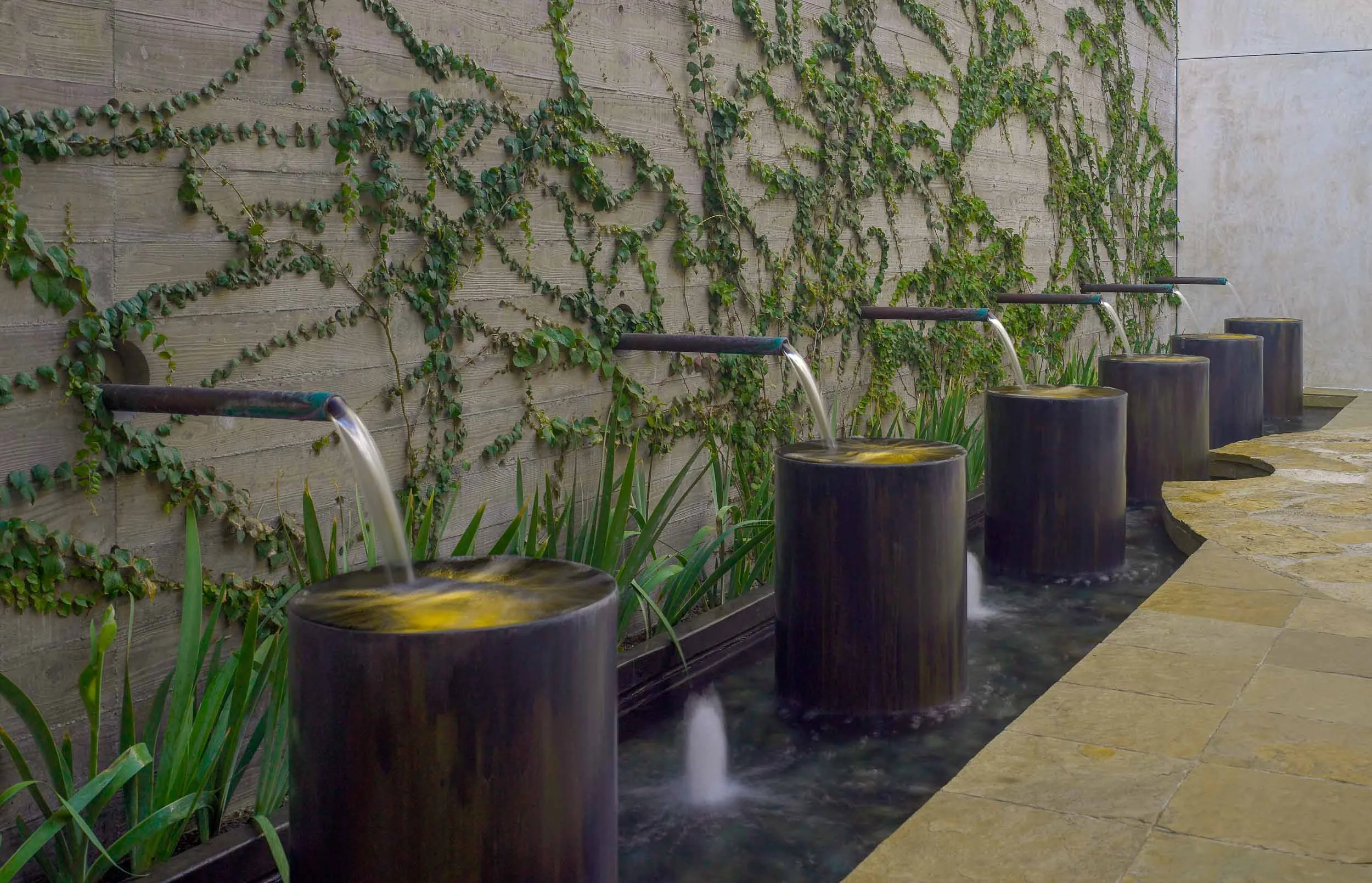 University of Santa Monica Garden Located on axis with the lobby hallway of the University of Santa Monica this garden patio is immediately visible upon entering the building. Pipes projecting out of the wall spill water into brass drums. Between each water drum, bubblers gurgle. Alternating sounds from the two water sources soothe the mind as they mask traffic noise from the nearby boulevard. Underwater lights within and between each drum animate the water’s movement and wash the vine covered wall. With each being controlled independently, the dual sources of lights and water create a variety of “scenes.” With water pipes off and lights within the drums on, the fountain becomes a serene scene of glowing circular planes of light. With bubblers on full, and water pipes flowing, the fountain changes from a quiet, contemplative focus to a highly animated experience. Flagstone circular paving focuses on the sculptural ‘icon’ of the University. The required exit stair has been redesigned and rebuilt in steel to become another element within the patio space. Olive trees, planted areas, and vines soften the hard surfaces.