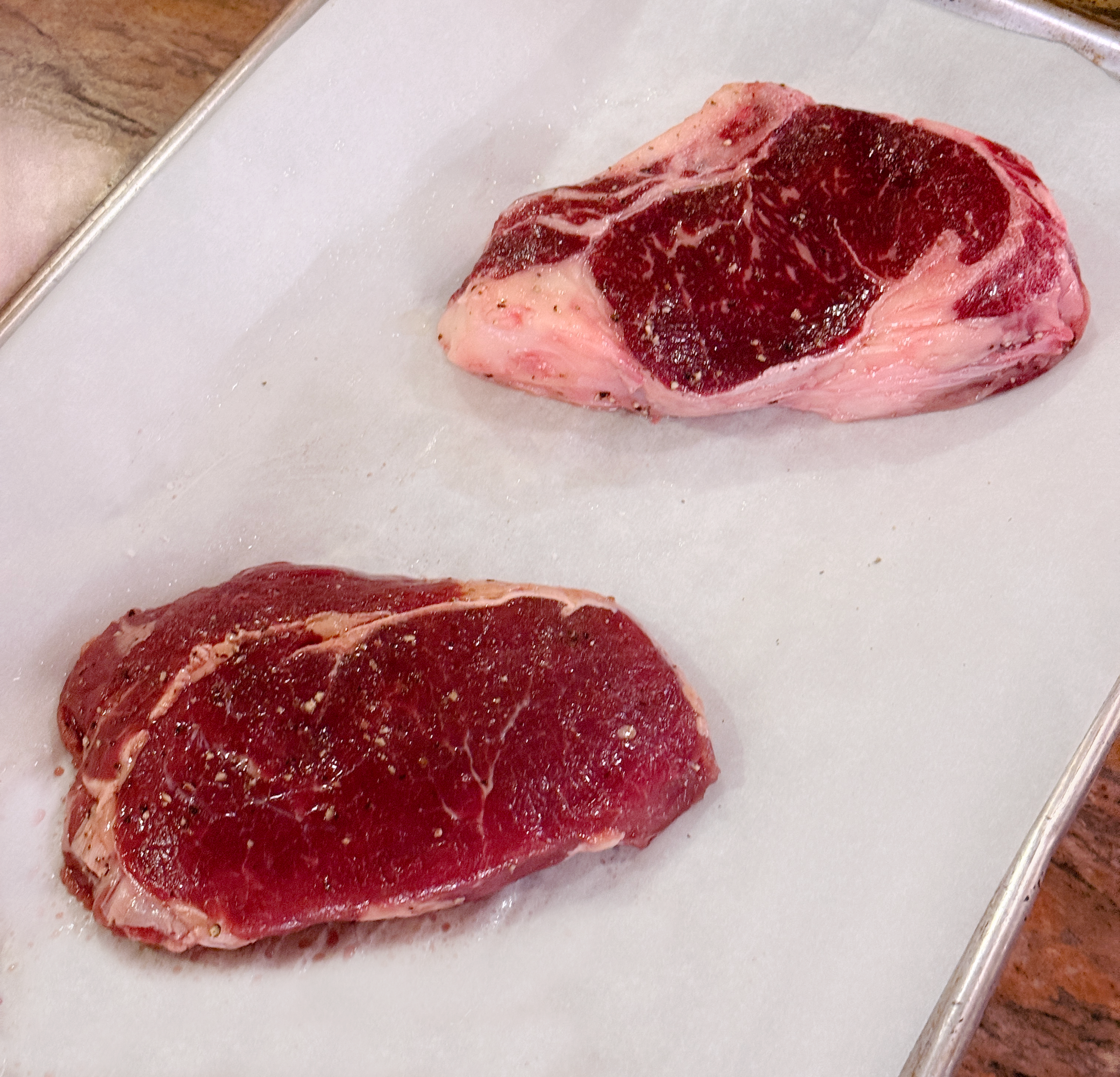 A photo of two raw, boneless, grass-fed ribeye steaks with salt and pepper on them, laying on top of a parchment covered pan.