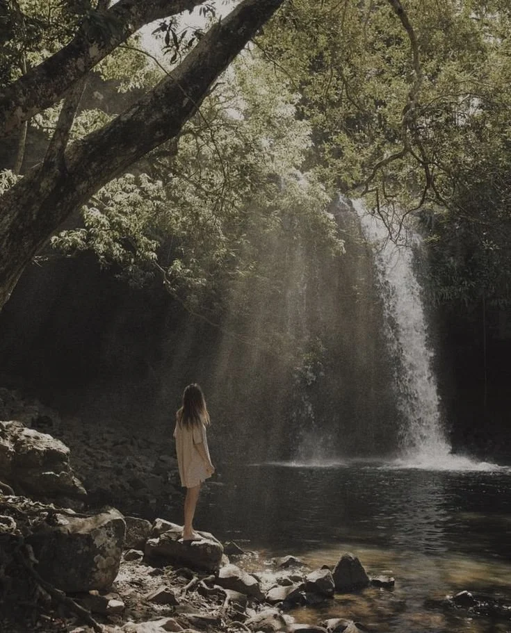 A young girl standing on rocks by a river, looking at a waterfall in a wooded area with sunlight filtering through the trees.
