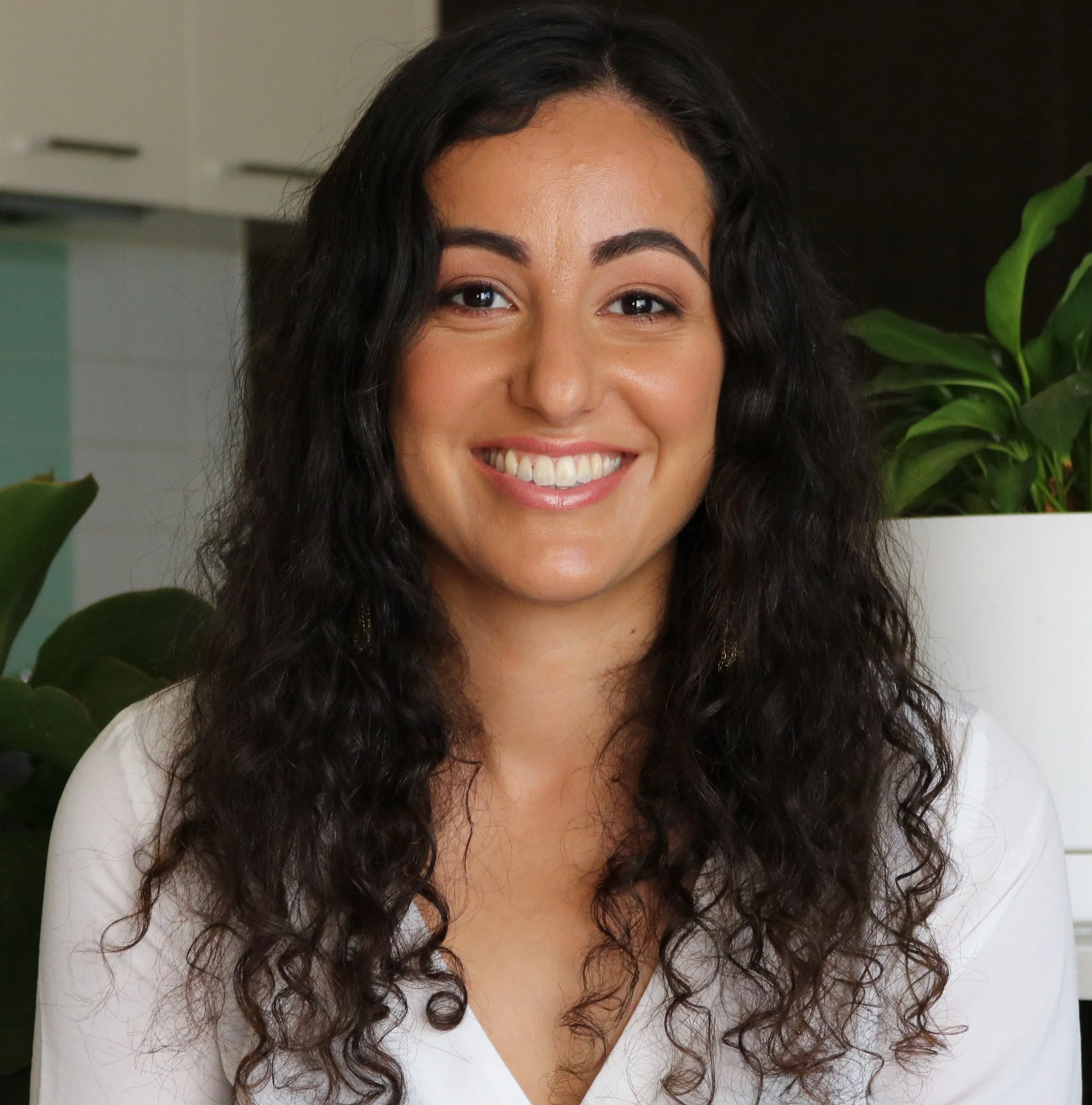 Smiling woman with long curly hair in a white shirt, standing indoors with houseplants in the background.