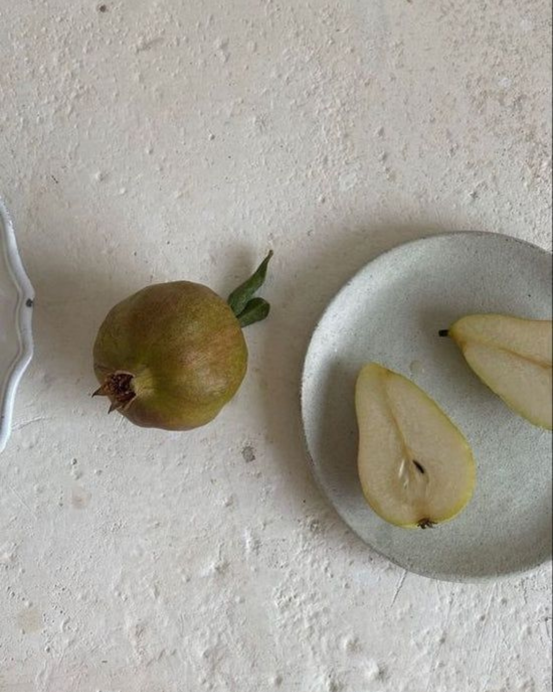 A whole greenish-brown pear with a stem and leaves next to a white plate holding two pear slices on a textured surface.
