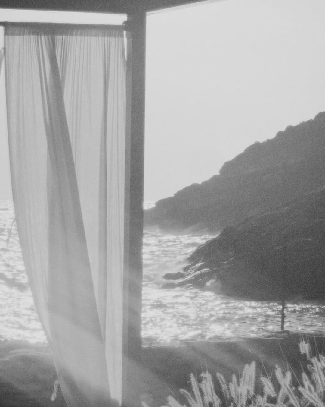 Beach scene viewed from inside a structure with sheer curtains, overlooking the ocean with rocks and a large canopy or umbrella.