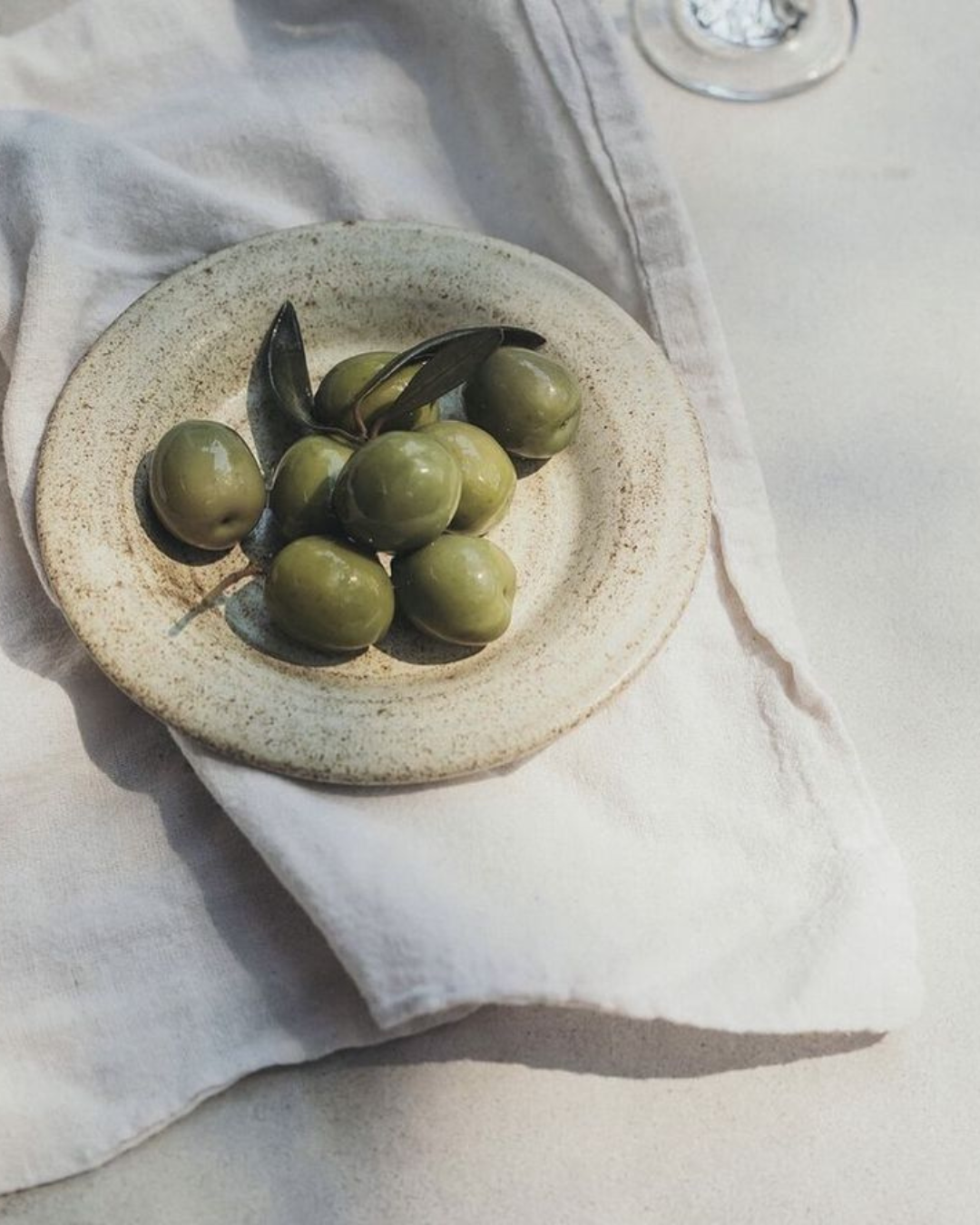 A small ceramic dish with green olives and a few dark leaves, placed on a white cloth.