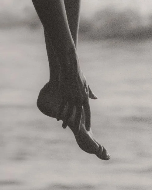 A black and white photo of a person's hand reaching down towards their toes, with the background blurred.
