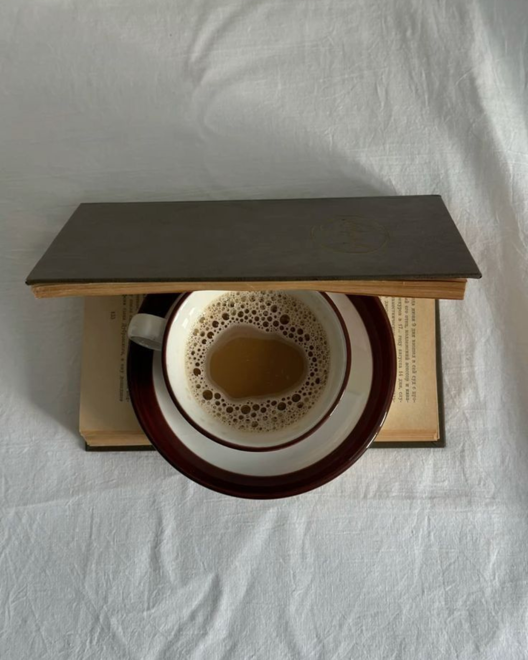 A cup of coffee on a saucer, resting on an open book, with an object resembling a closed book or wooden cover on top, viewed from above.