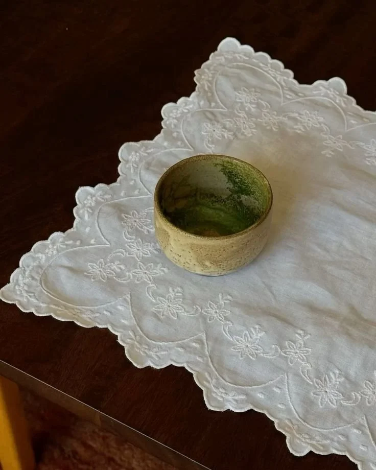 A ceramic bowl with a mossy green and beige glaze, resting on a white embroidered tablecloth with scalloped edges and floral embroidery, on a dark wooden table.