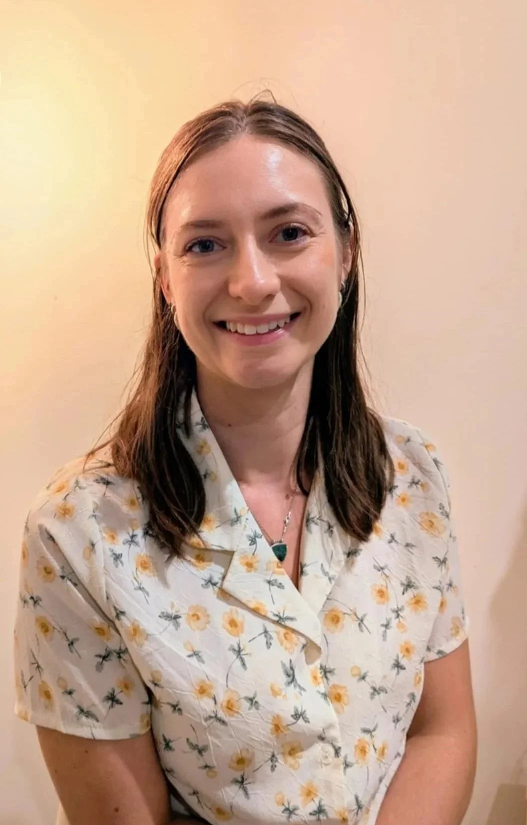 A smiling woman with shoulder-length brown hair, wearing a light-colored floral blouse and a necklace, standing against a plain beige wall.