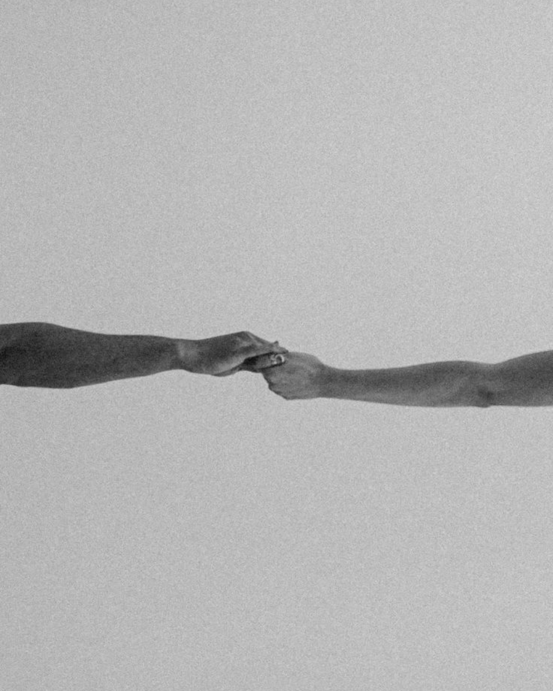 Two hands reaching out and holding each other's fingers against a plain light gray background.
