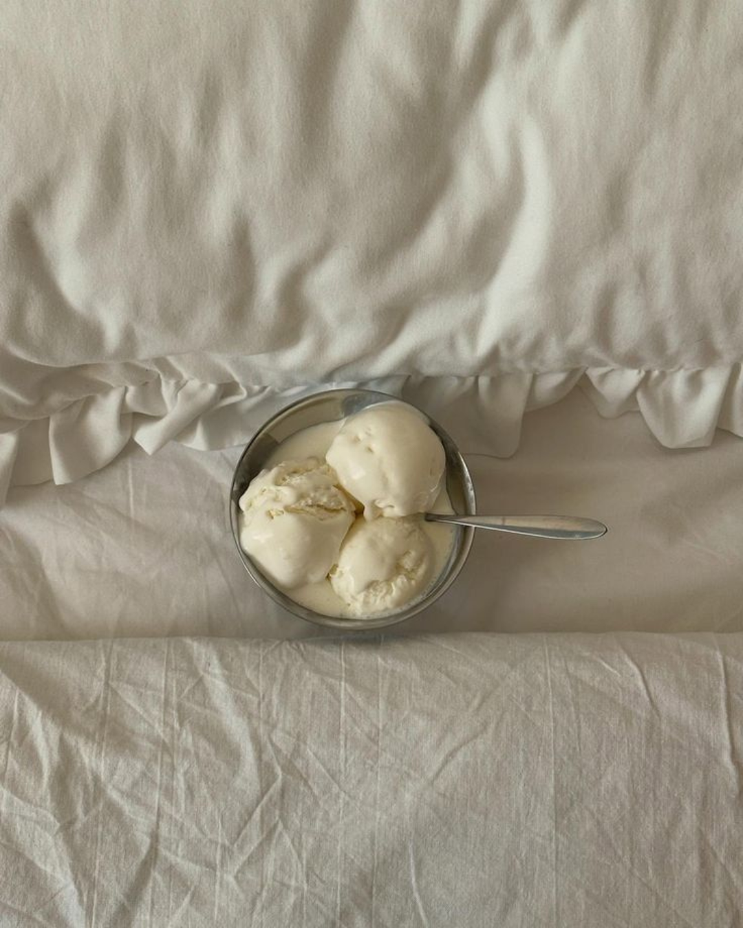 A bowl of three scoops of vanilla ice cream with a spoon, placed on a white fabric surface with a crumpled sheet or blanket overhead.