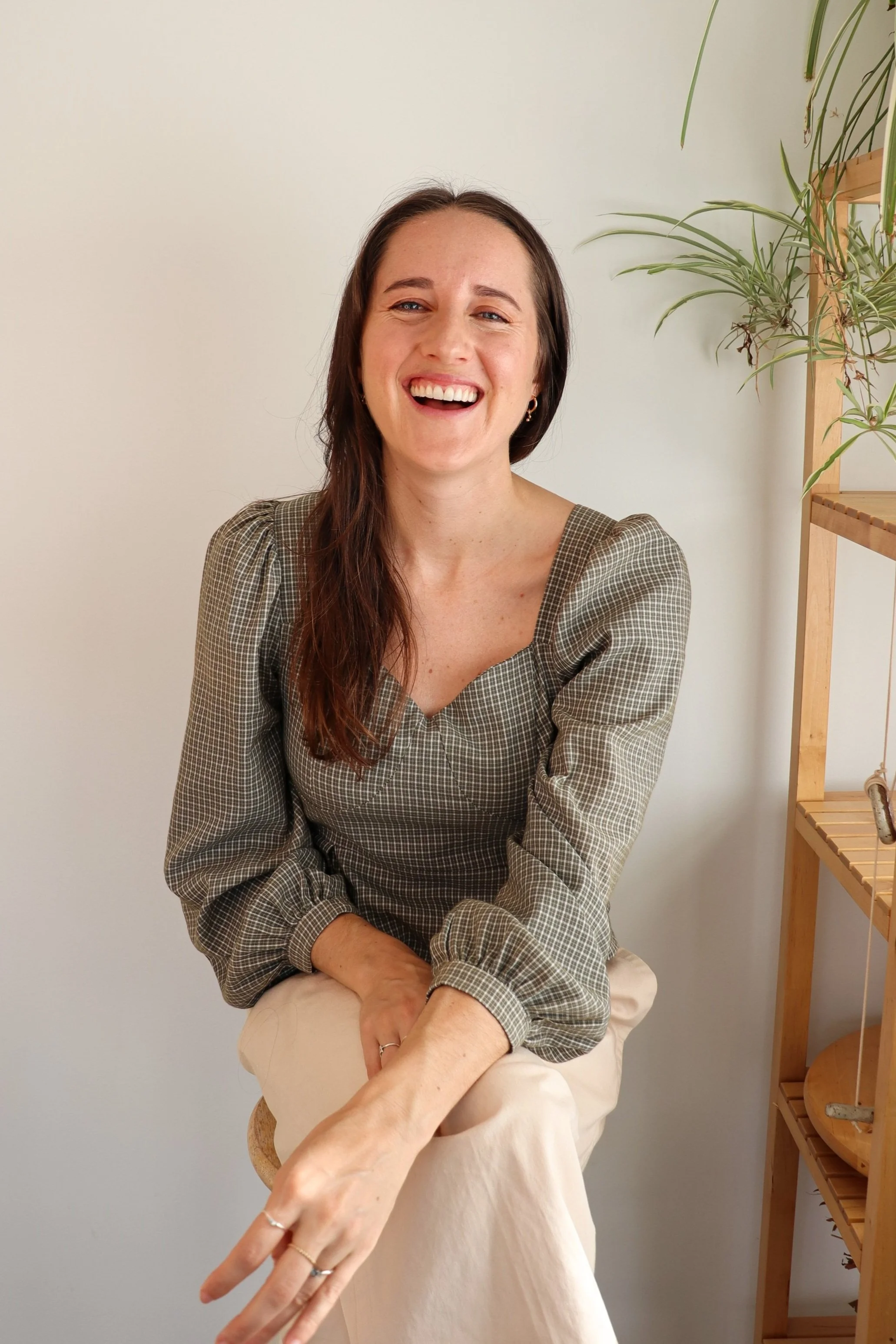 Woman smiling, sitting on a stool, wearing a checkered blouse and light pants, with a plant on a wooden shelf in the background.