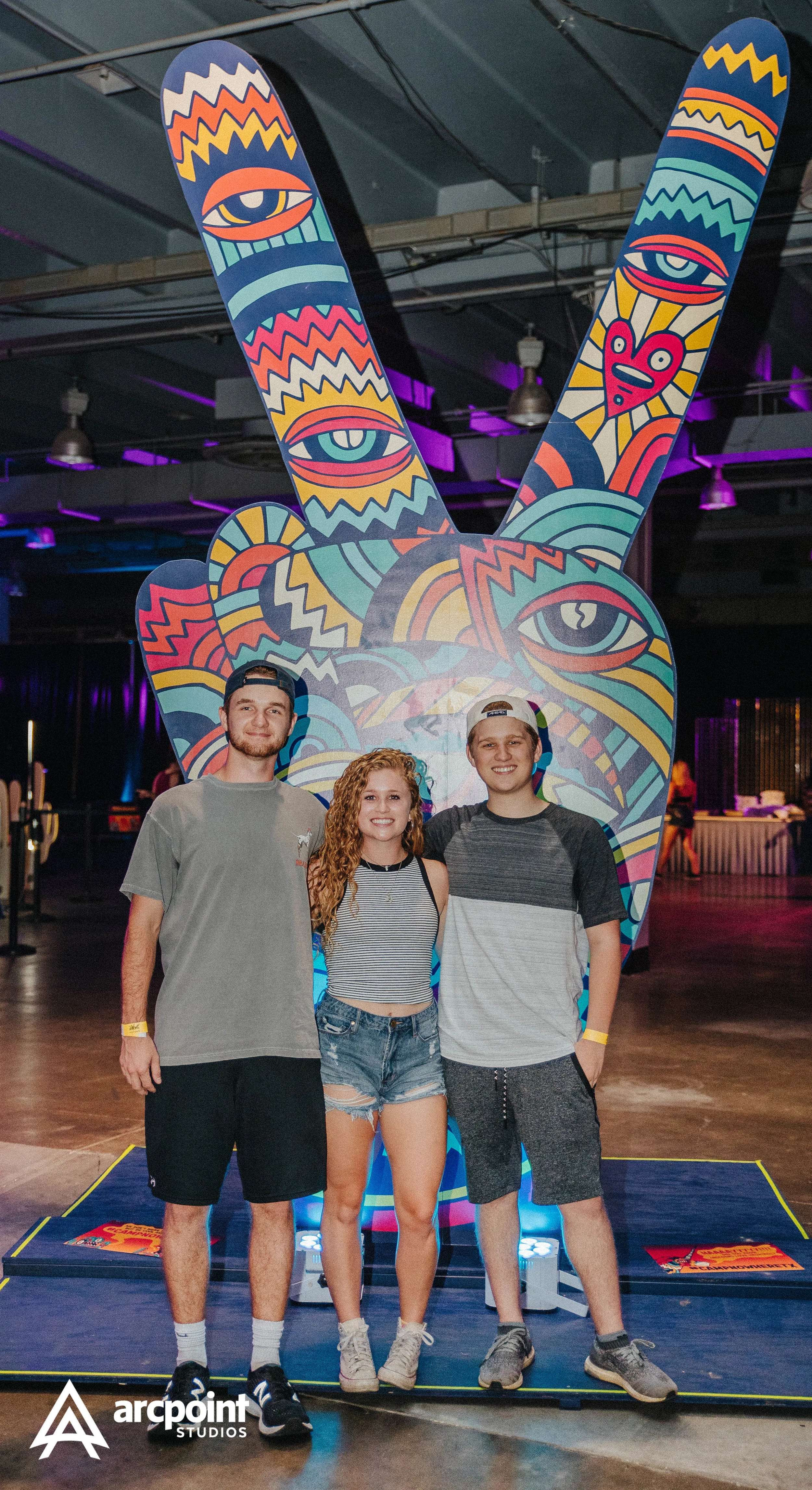 Three young people, two males and one female, standing in front of a colorful artistic sculpture of a hand making a peace sign, with eye and face imagery. They are inside a dimly lit venue with purple and blue lighting.