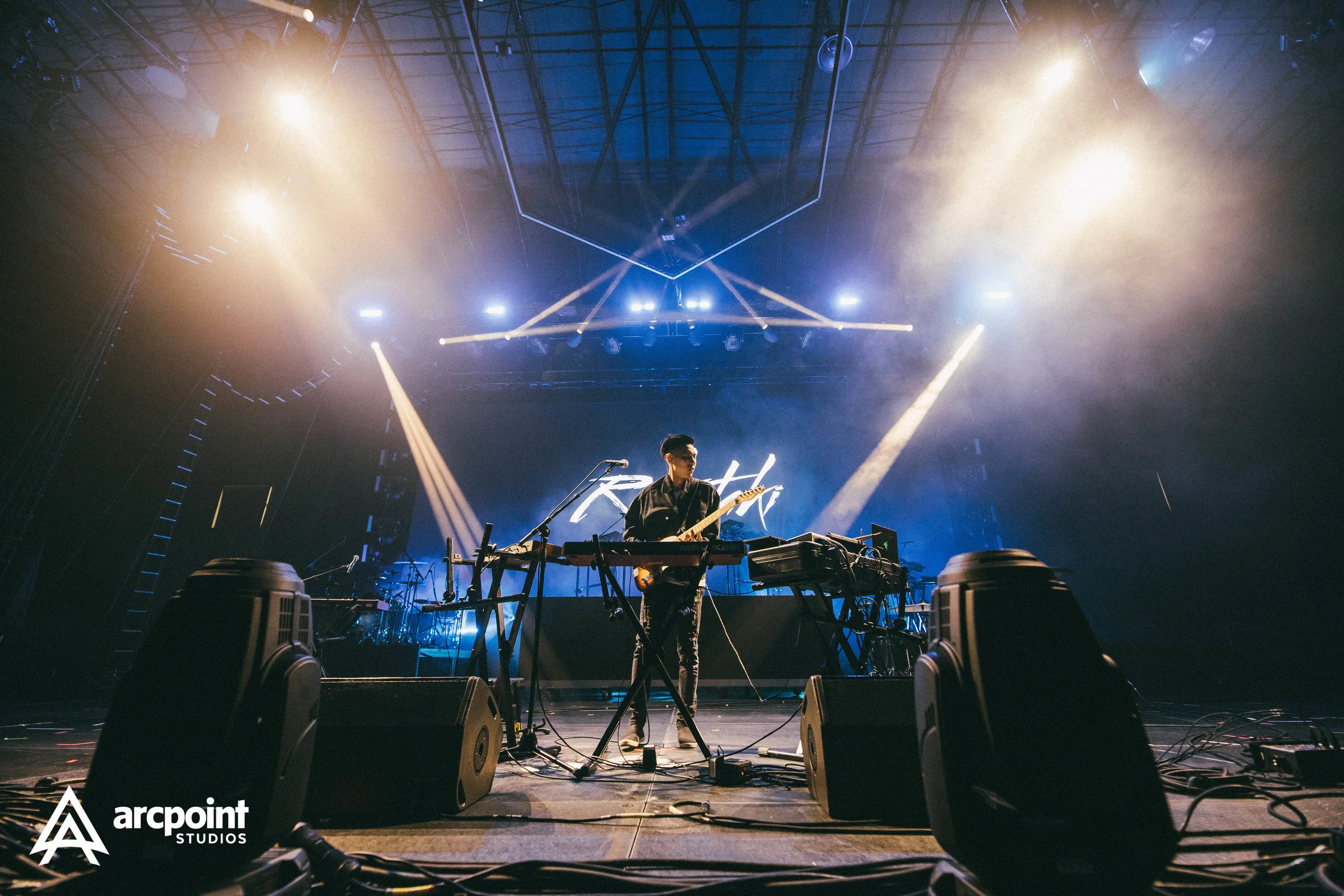 A musician playing a keyboard and guitar on stage during a live concert with colorful lighting and smoke effects.