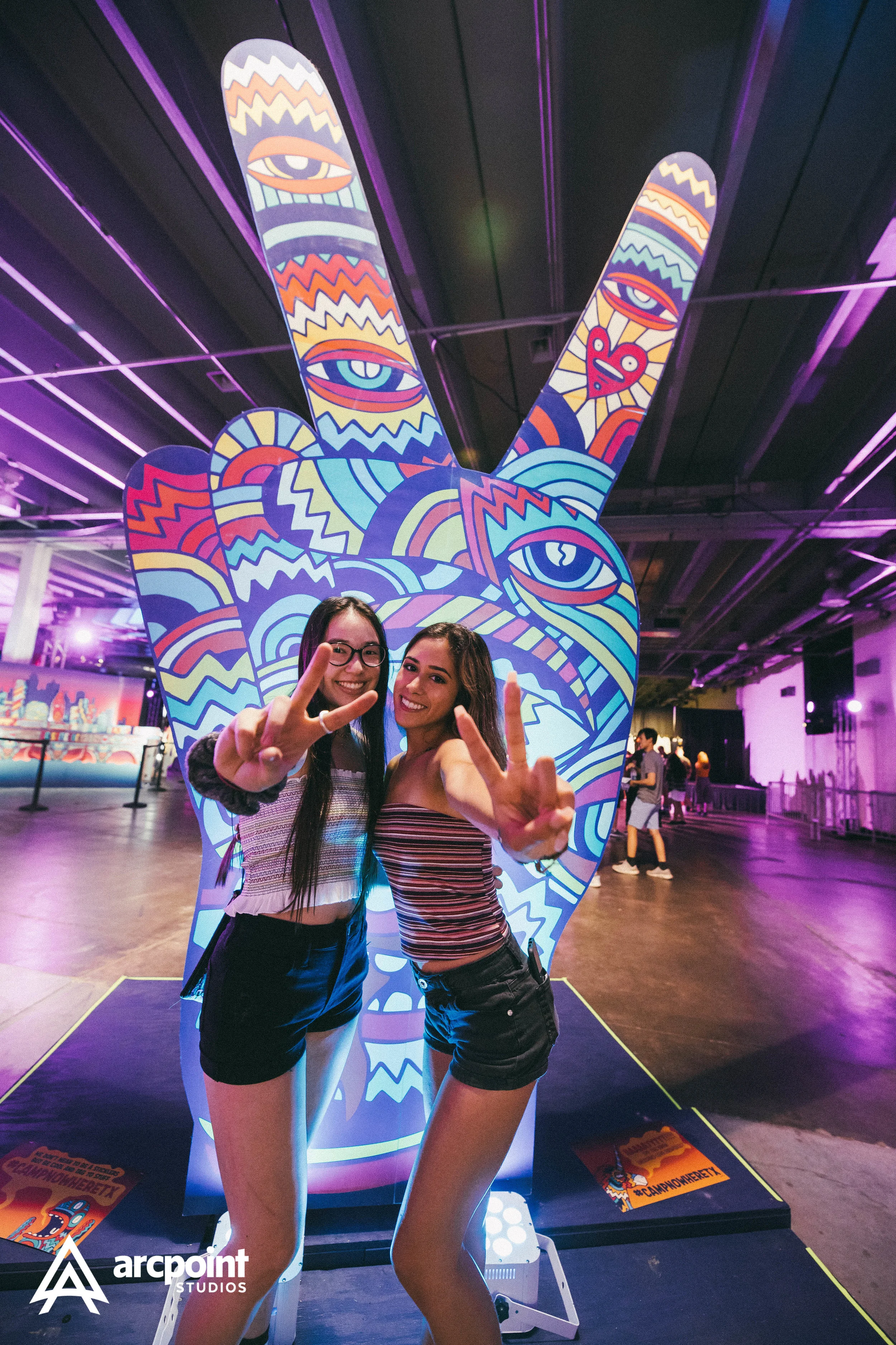 Two women smiling and making peace signs in front of a colorful, graffiti-style peace sign at an indoor event with purple lighting.