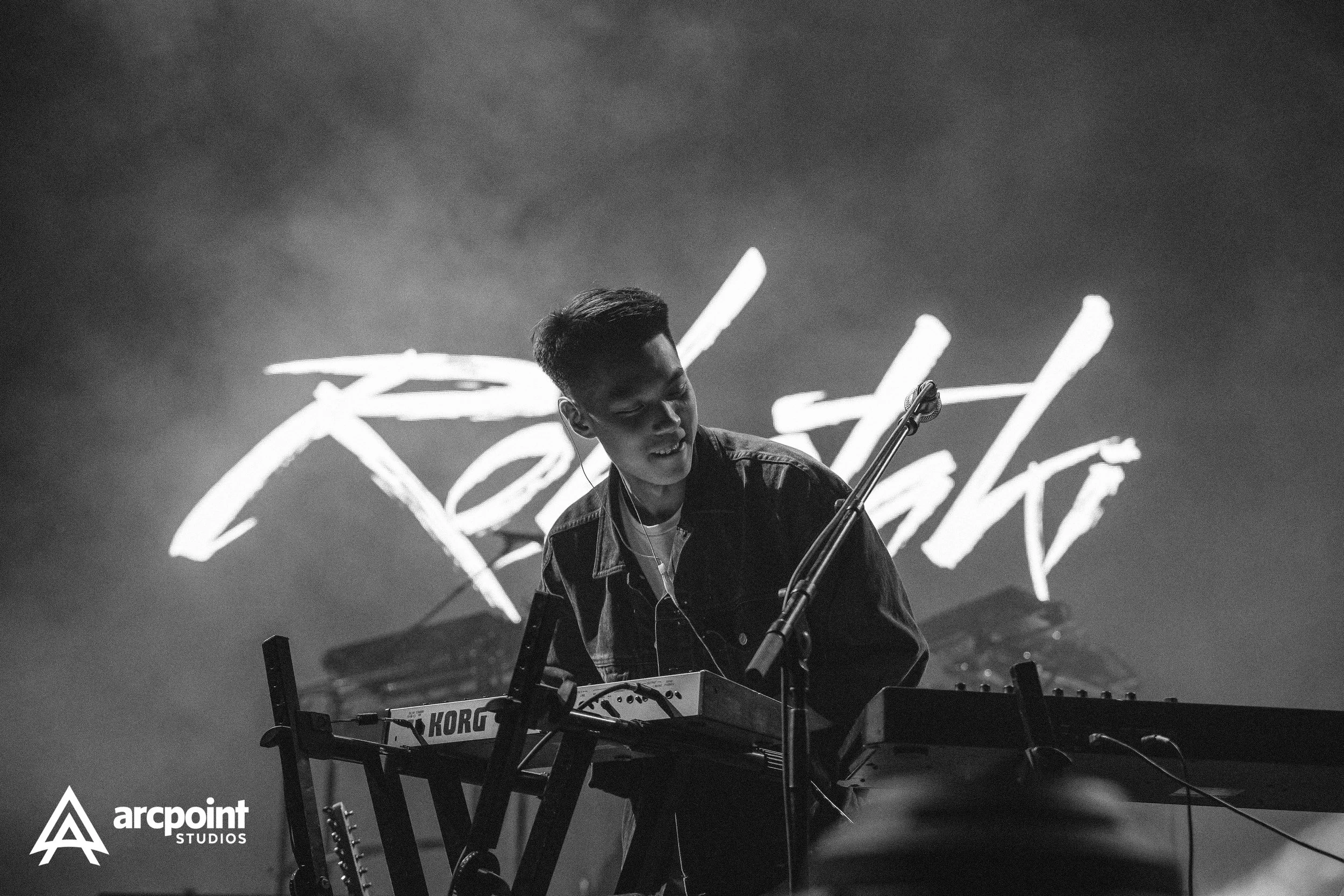 A musician performing live on stage, playing instruments with a backdrop displaying the word "Fearless" in neon lights, in black and white.
