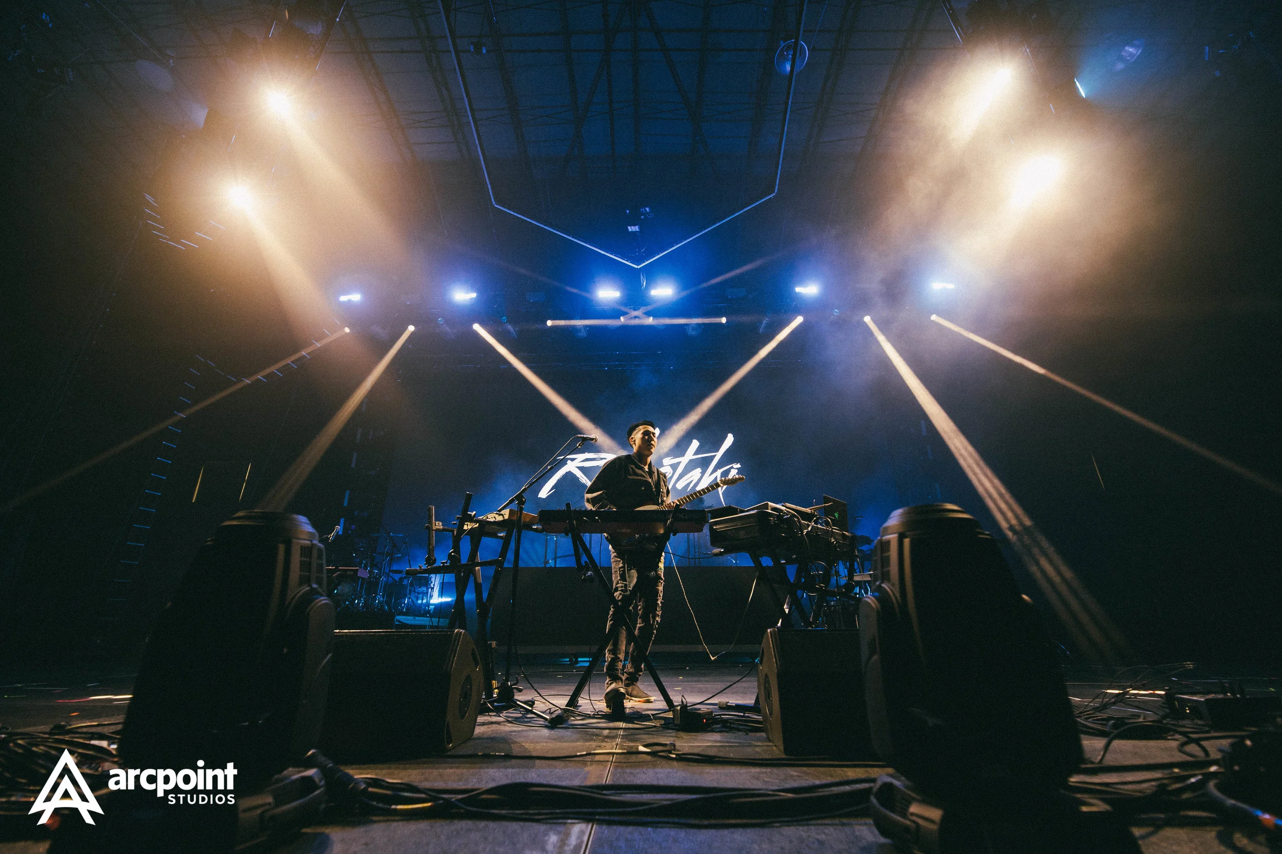 A musician stands on a stage with keyboards and electronic equipment, illuminated by colorful stage lights, performing at a concert or music event with the band name 'Kasti' displayed on a screen behind.