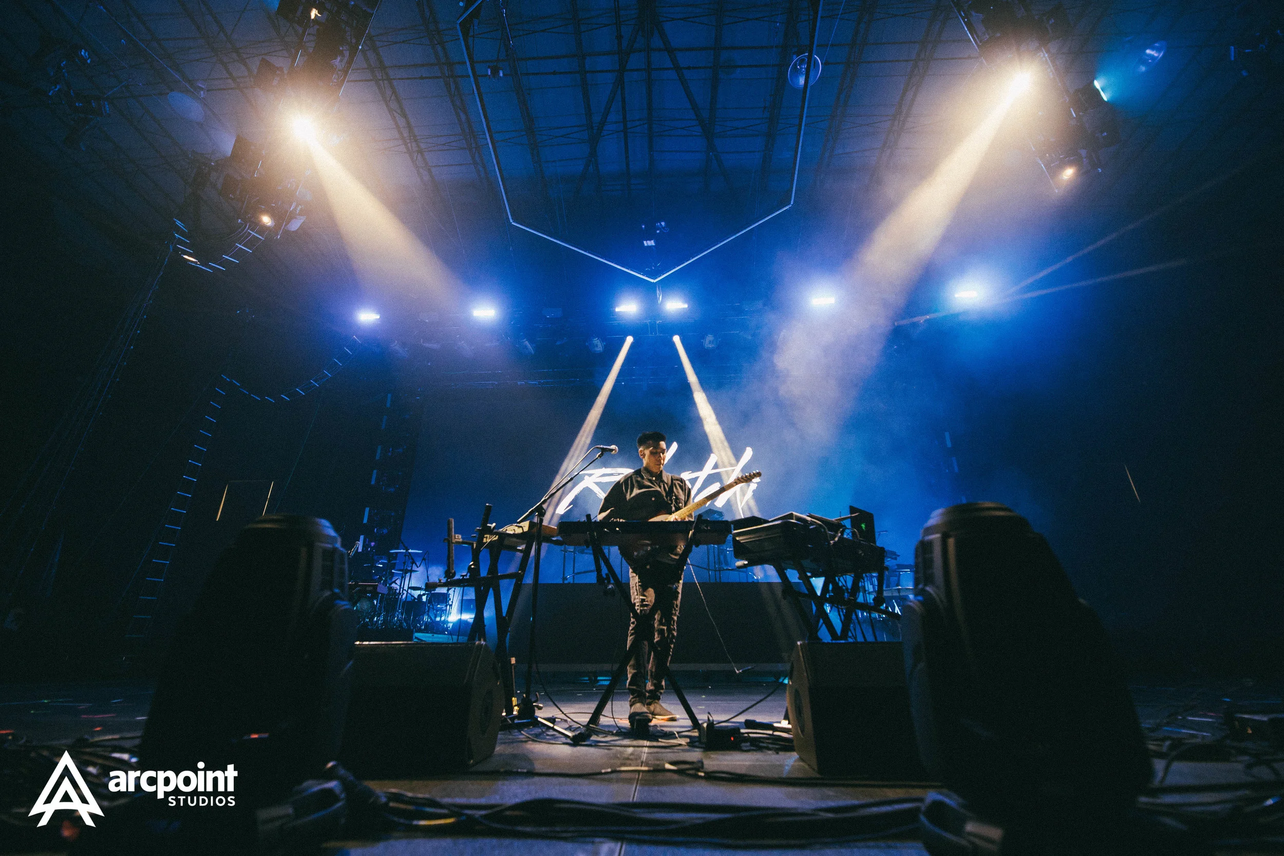 A musician performing on stage with electronic music equipment, illuminated by bright stage lights, with fog effects, in a concert setting.