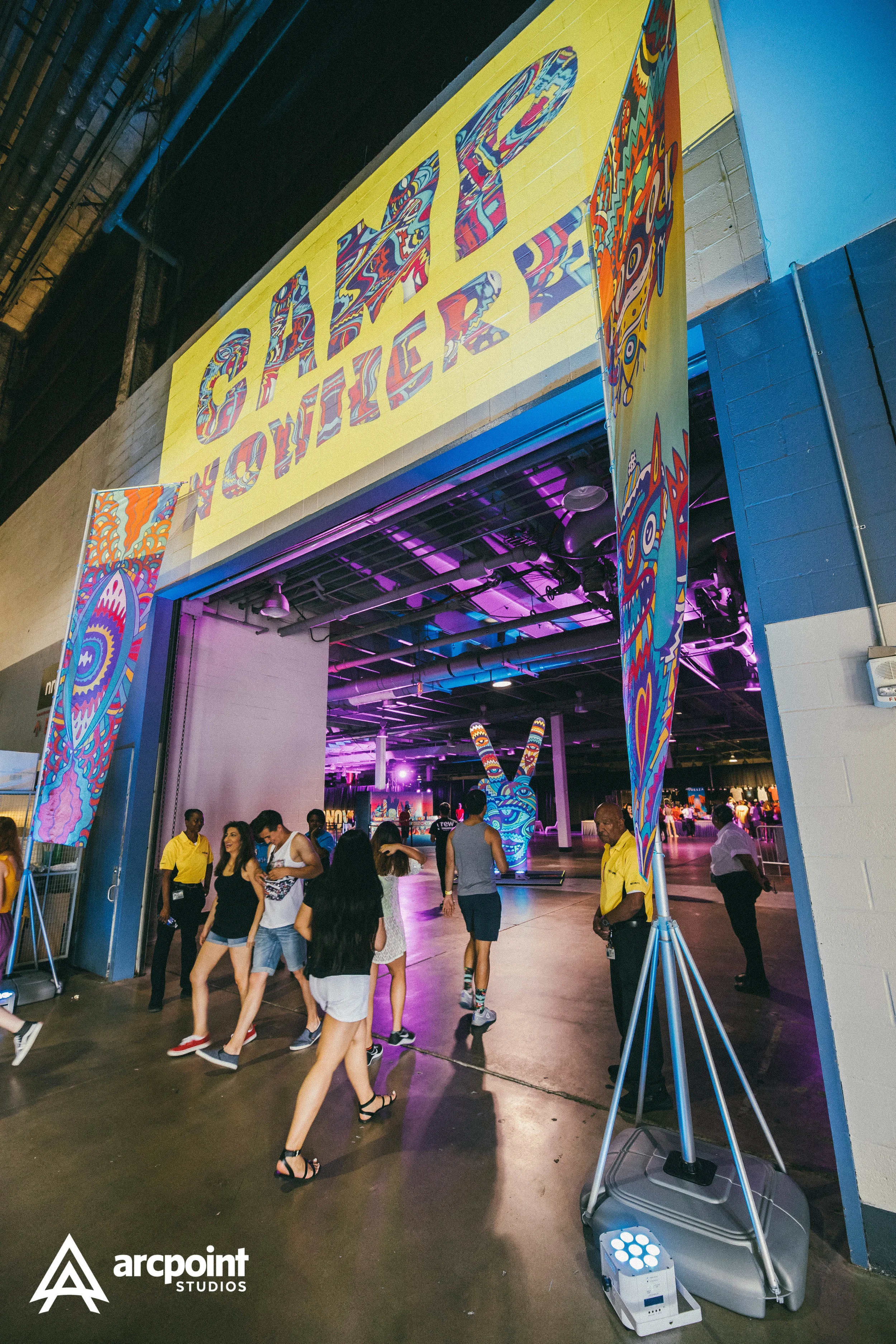 People entering the California Welcome Center at a bright, colorful event with psychedelic art and large Peace Hand and Eye symbols, illuminated with purple and pink lighting.