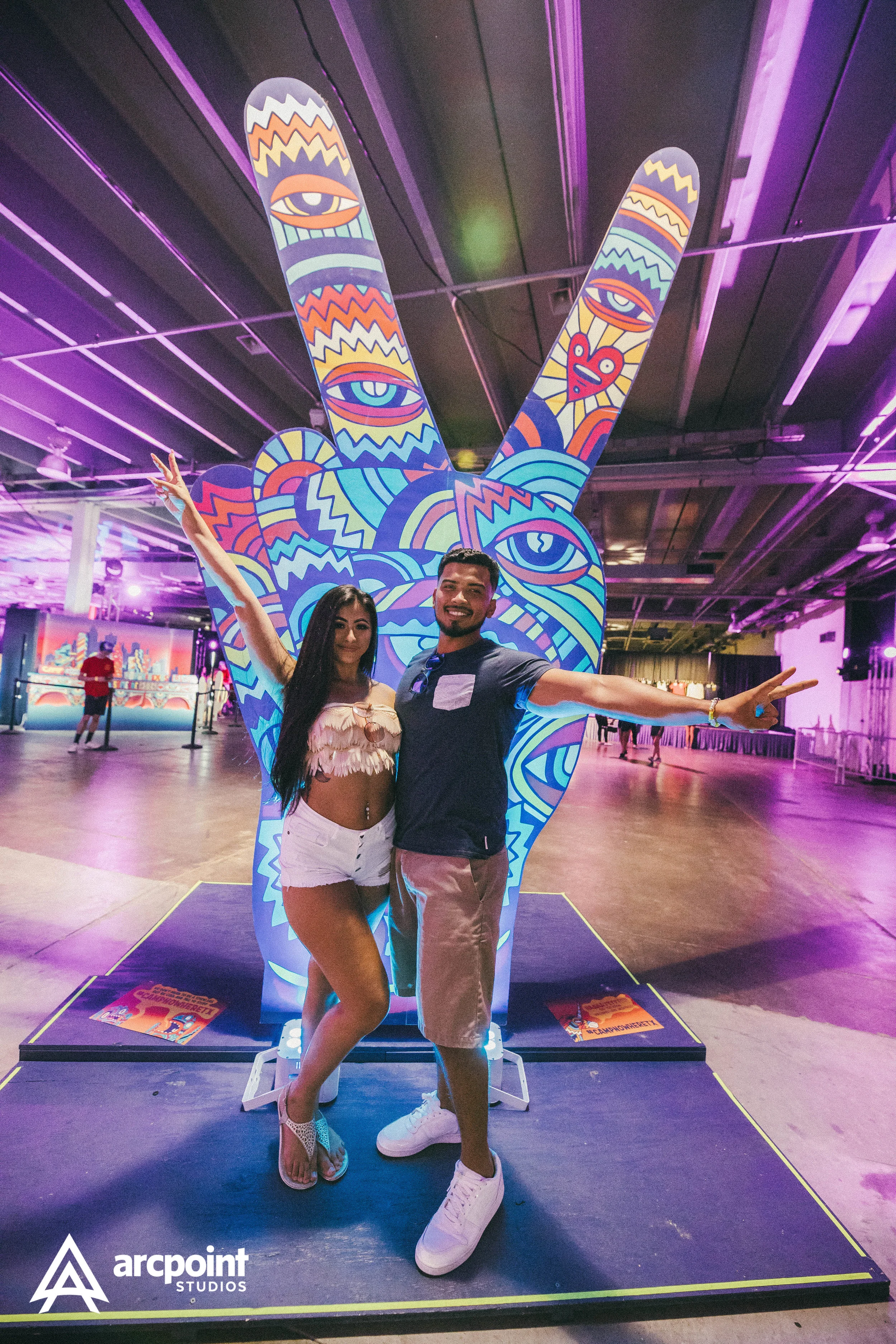 A man and a woman posing in front of a colorful, psychedelic peace sign sculpture at an indoor event, with purple lighting overhead.