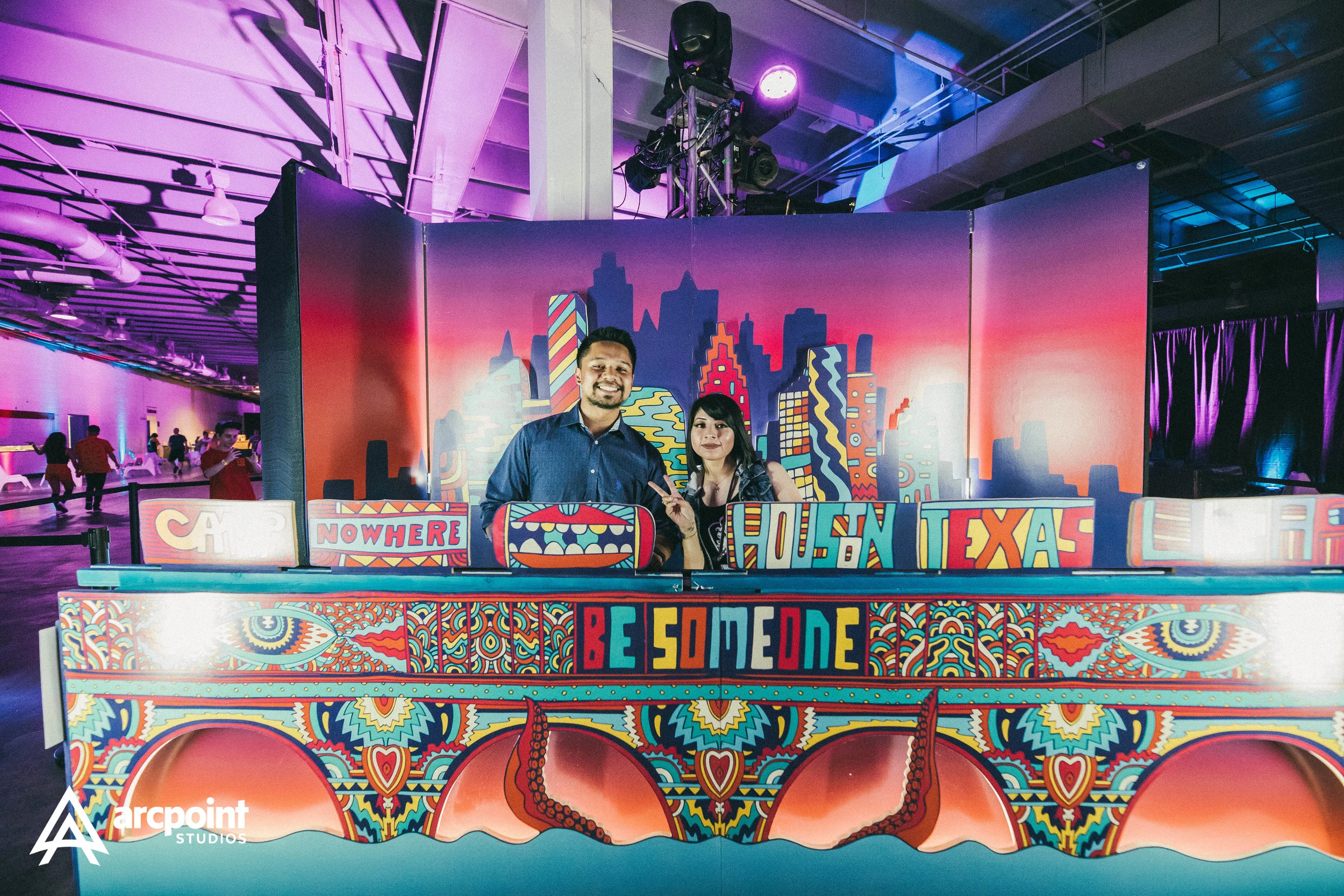 Two people smiling behind a colorful, graffiti-style bar with a cityscape banner in the background and purple lighting in an indoor event space.