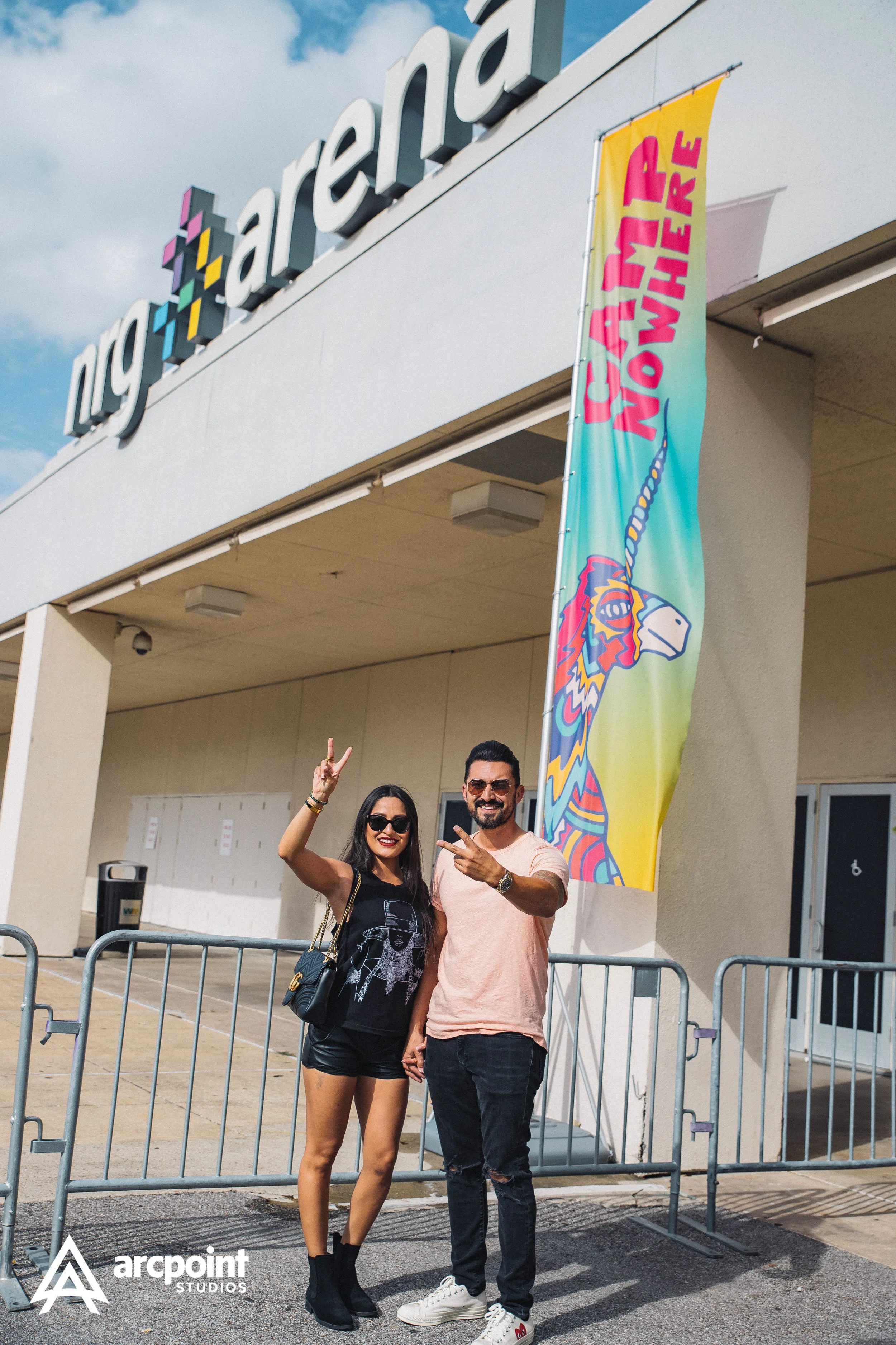 Two smiling people, a woman and a man, stand in front of a building with a large sign that reads 'ng arena'. They are making peace signs and holding hands. The woman is wearing sunglasses, a black T-shirt, shorts, and boots. The man is wearing sungla