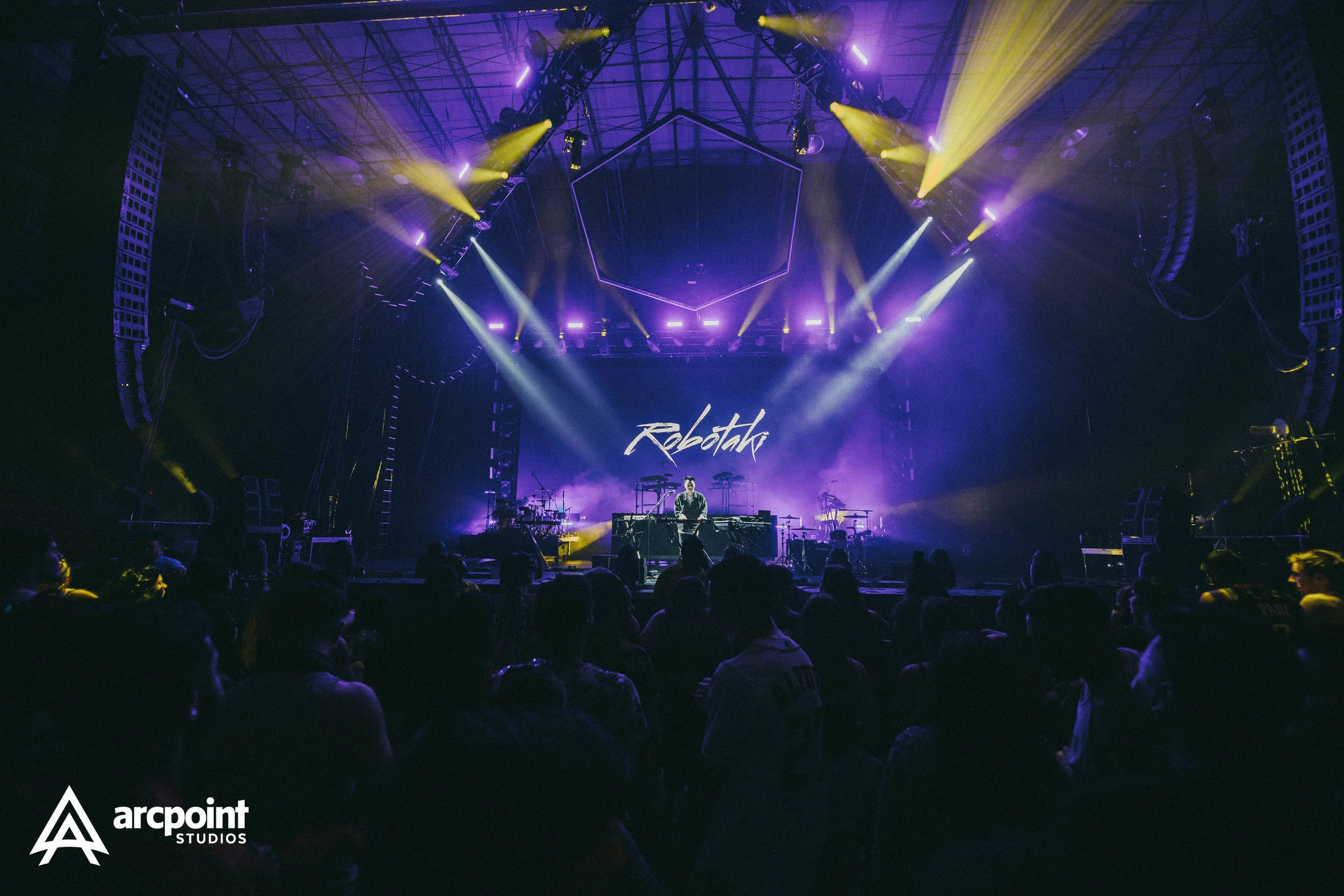 Concert stage with purple and yellow lighting, DJ performing at a music festival called Robatki, with an audience watching. Logo of arcpoint studios in the bottom left corner.