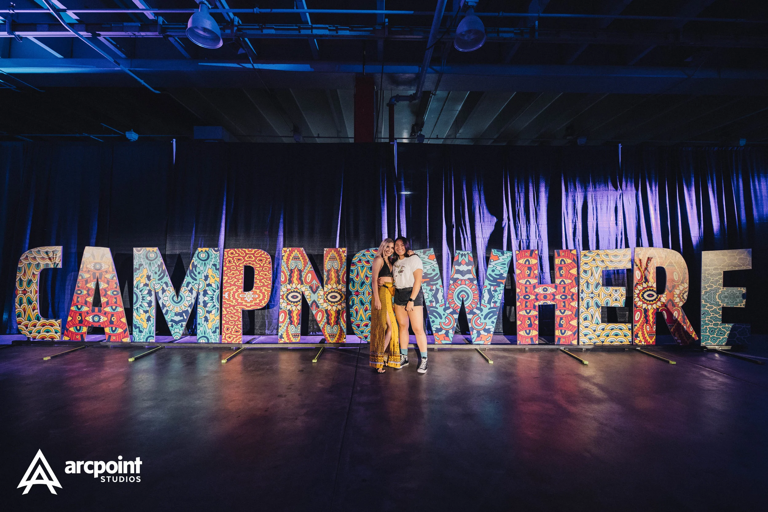Two young women standing in front of large, colorful, patterned letters spelling 'CAMP H'ER' on a stage with a dark curtain backdrop at a studio event.
