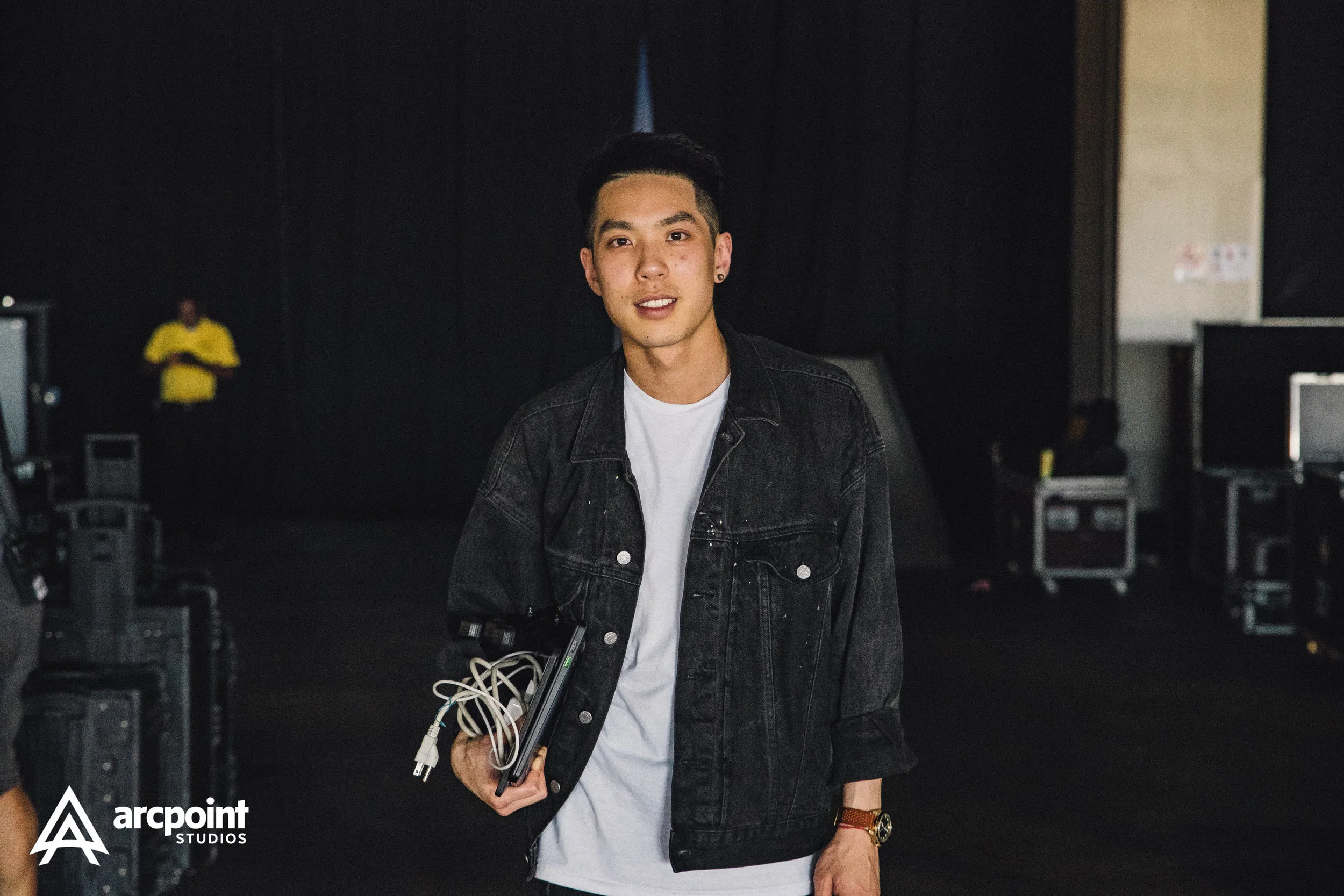 A young man standing in a dark room, holding a laptop with cables, wearing a black denim jacket, white t-shirt, and watch, with equipment and a person in a yellow shirt in the background.