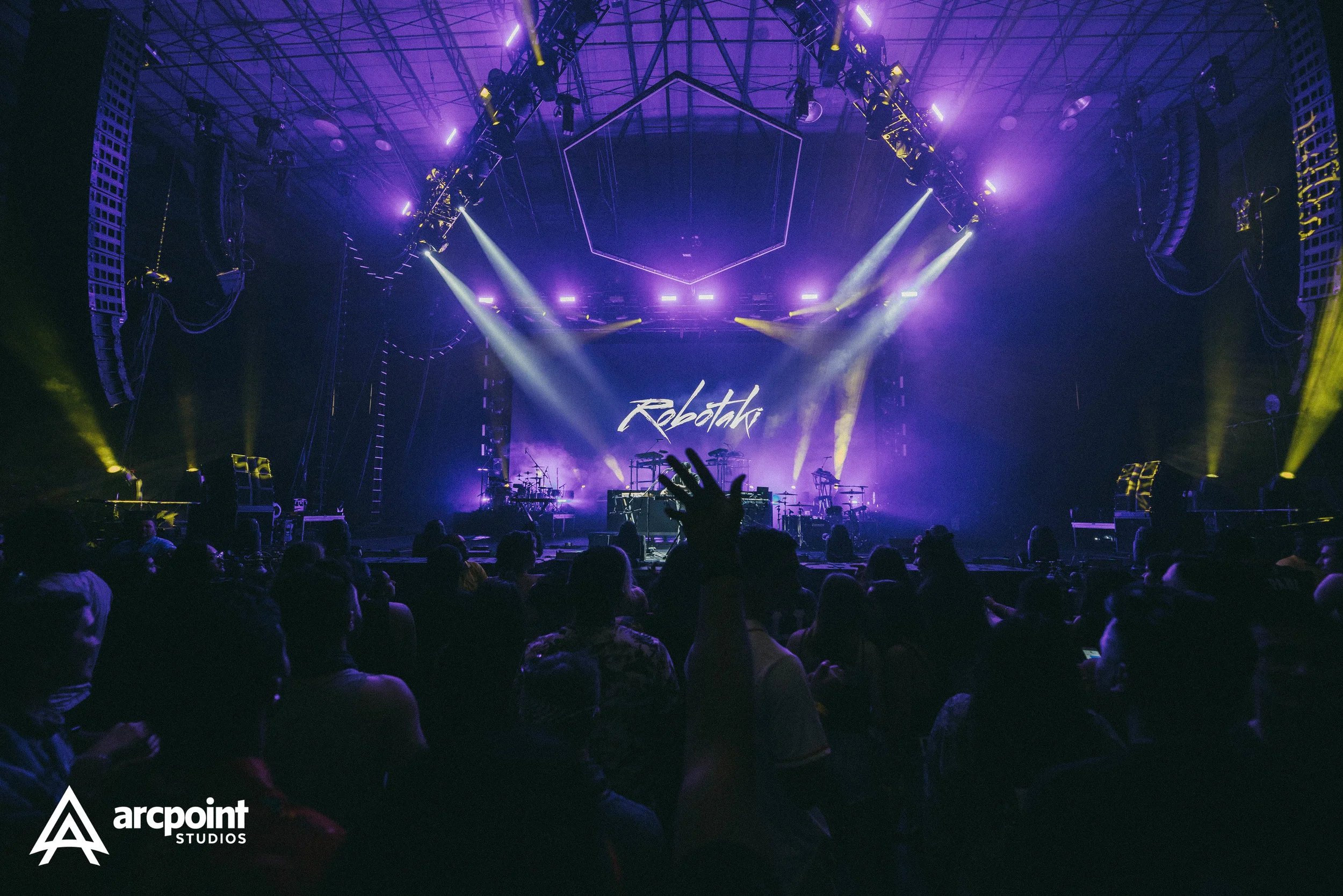 A concert stage with purple and yellow lighting, musical instruments, and the word "Reboot" displayed on a large screen. Audience members are seated in front of the stage, with one person raising their hand.