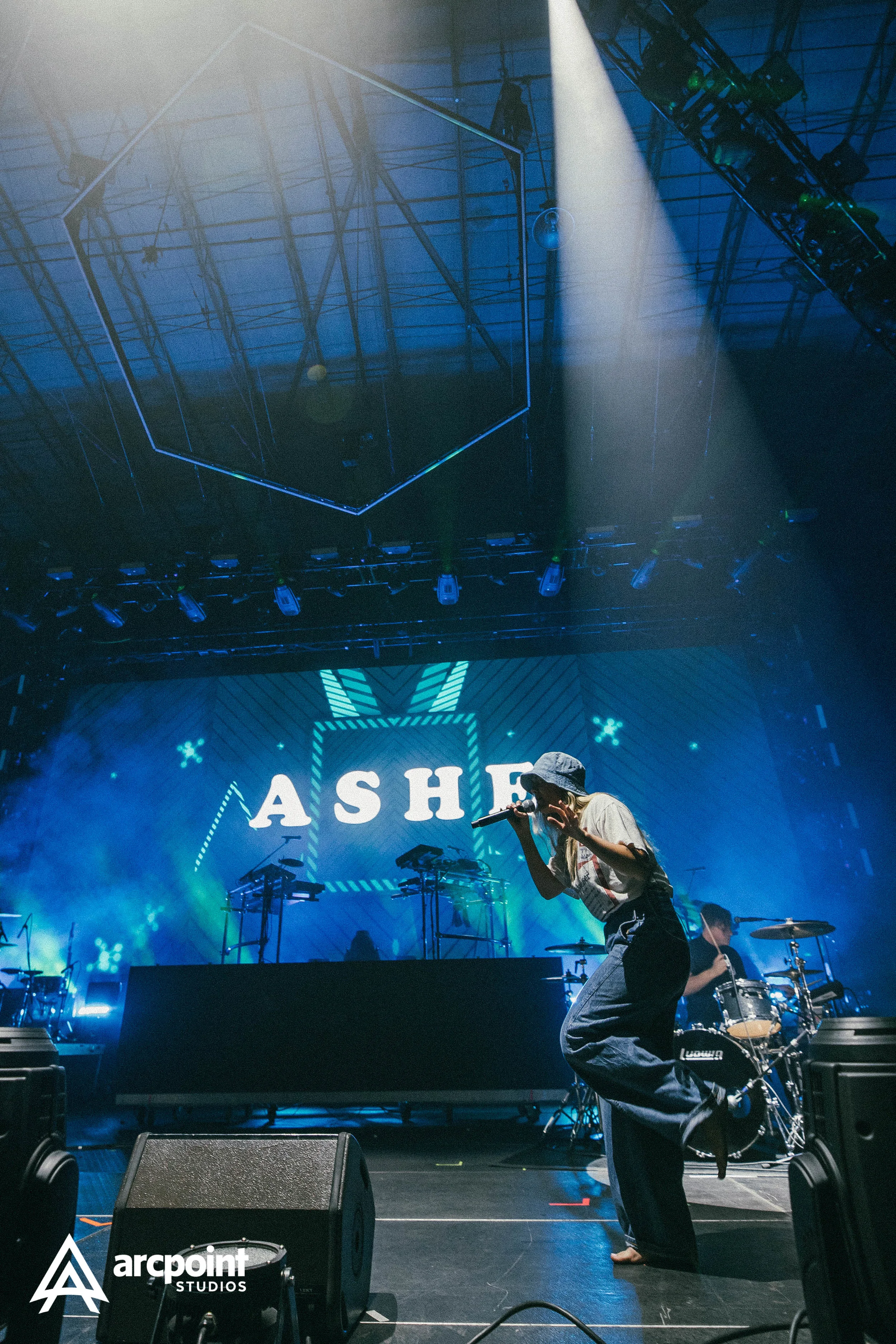 Performer wearing a hat and casual clothes singing into a microphone on stage with drum set behind, with a large screen in the background displaying the word 'ASHE' and colorful stage lights.