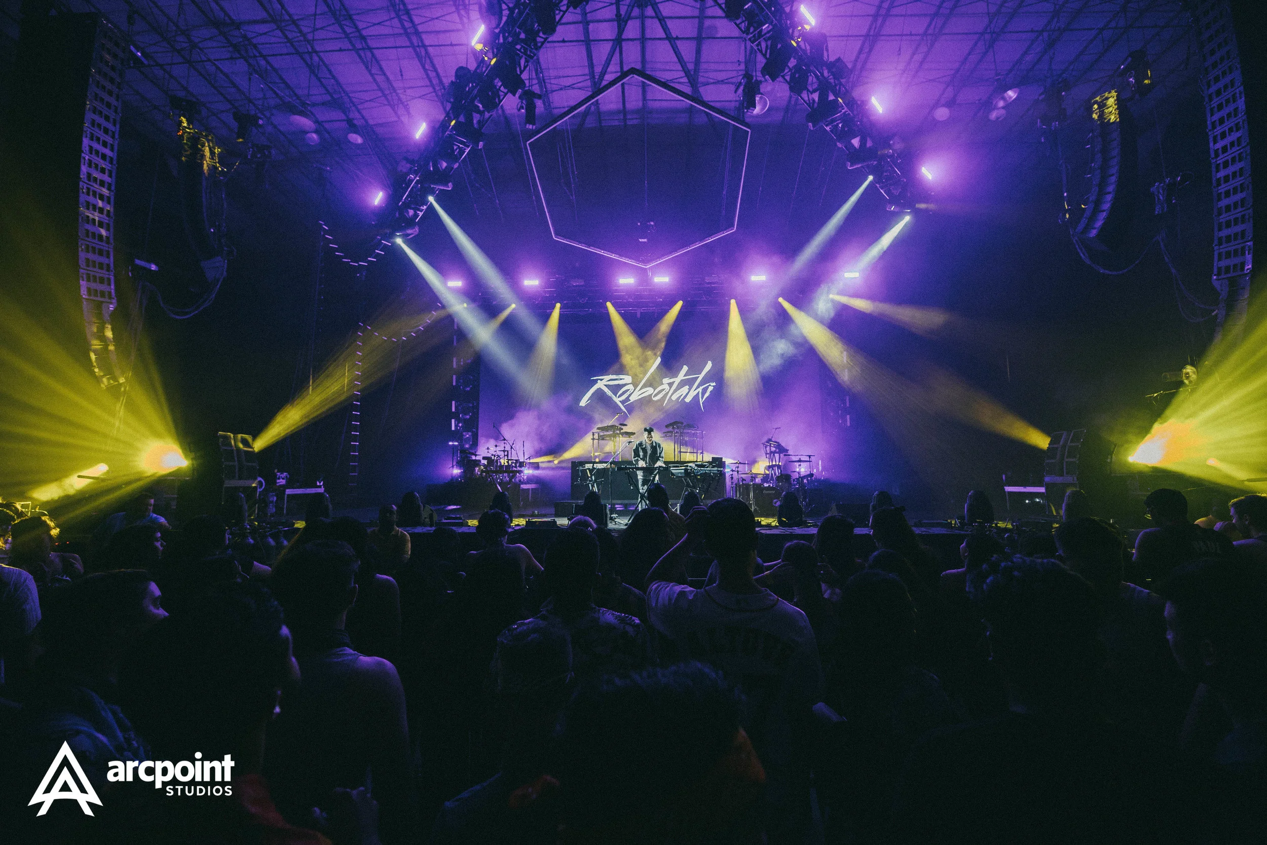 Concert stage with bright yellow, purple, and white lighting, a DJ or performer at the center with a backdrop displaying the name 'Robothk', and an audience in the foreground. The logo for 'arcpöint STUDIOS' is in the bottom left corner.