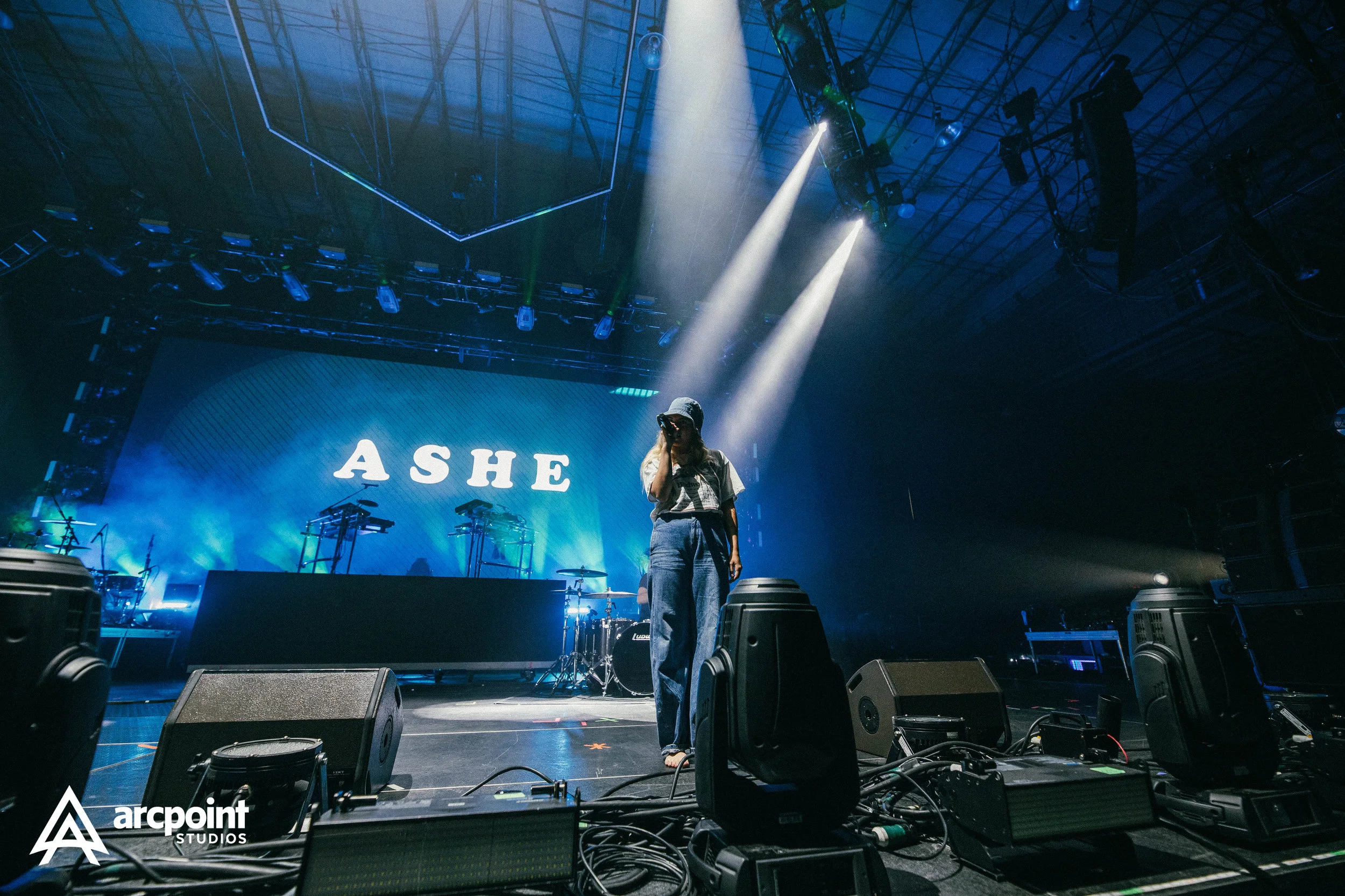 A person on stage with a microphone, standing in front of a large screen displaying the name ASHE, during a concert or performance, with stage lights and equipment around.