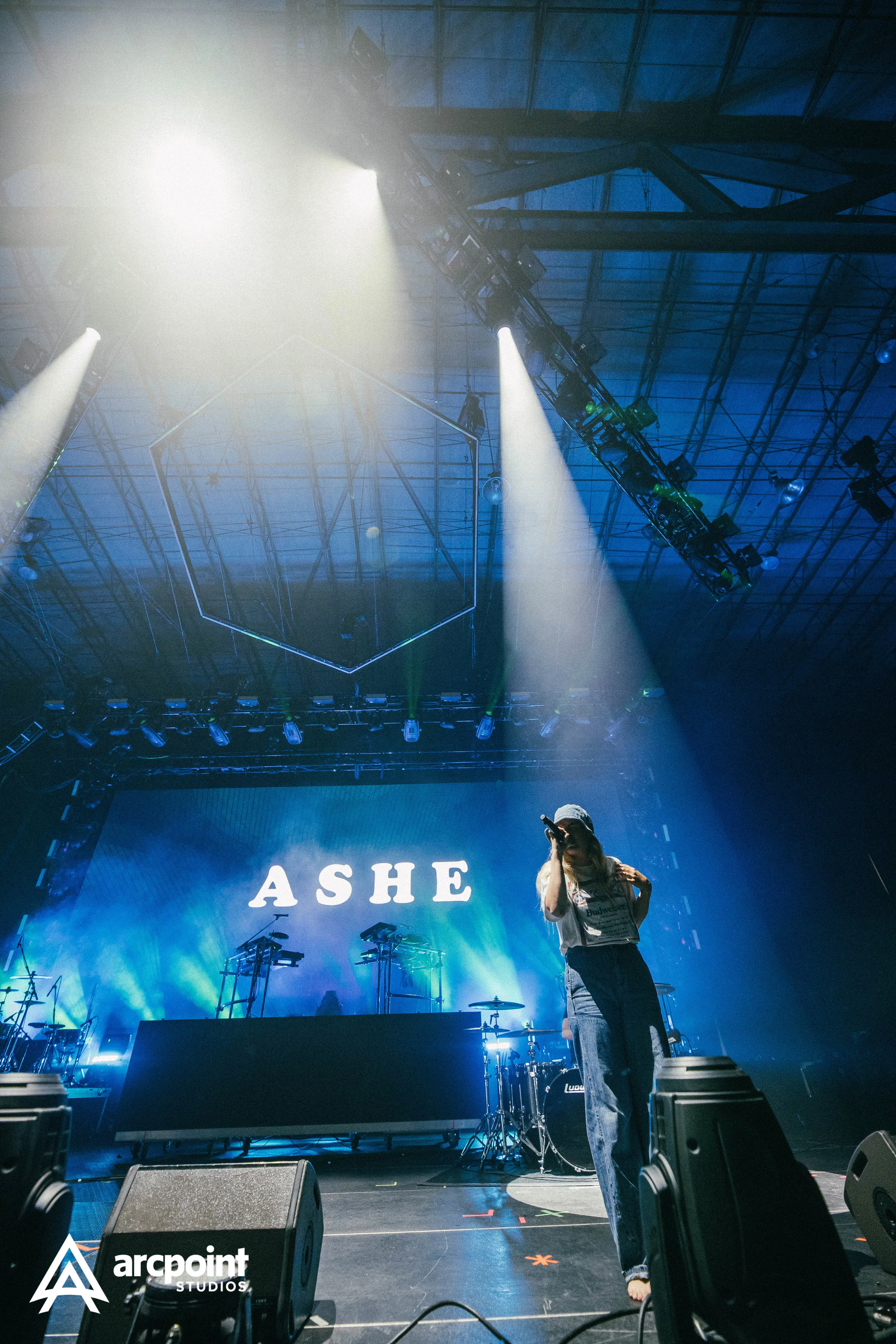 A performer on stage at a concert, with a bright spotlight shining from above, and the name "ASHE" displayed on a large screen behind them.