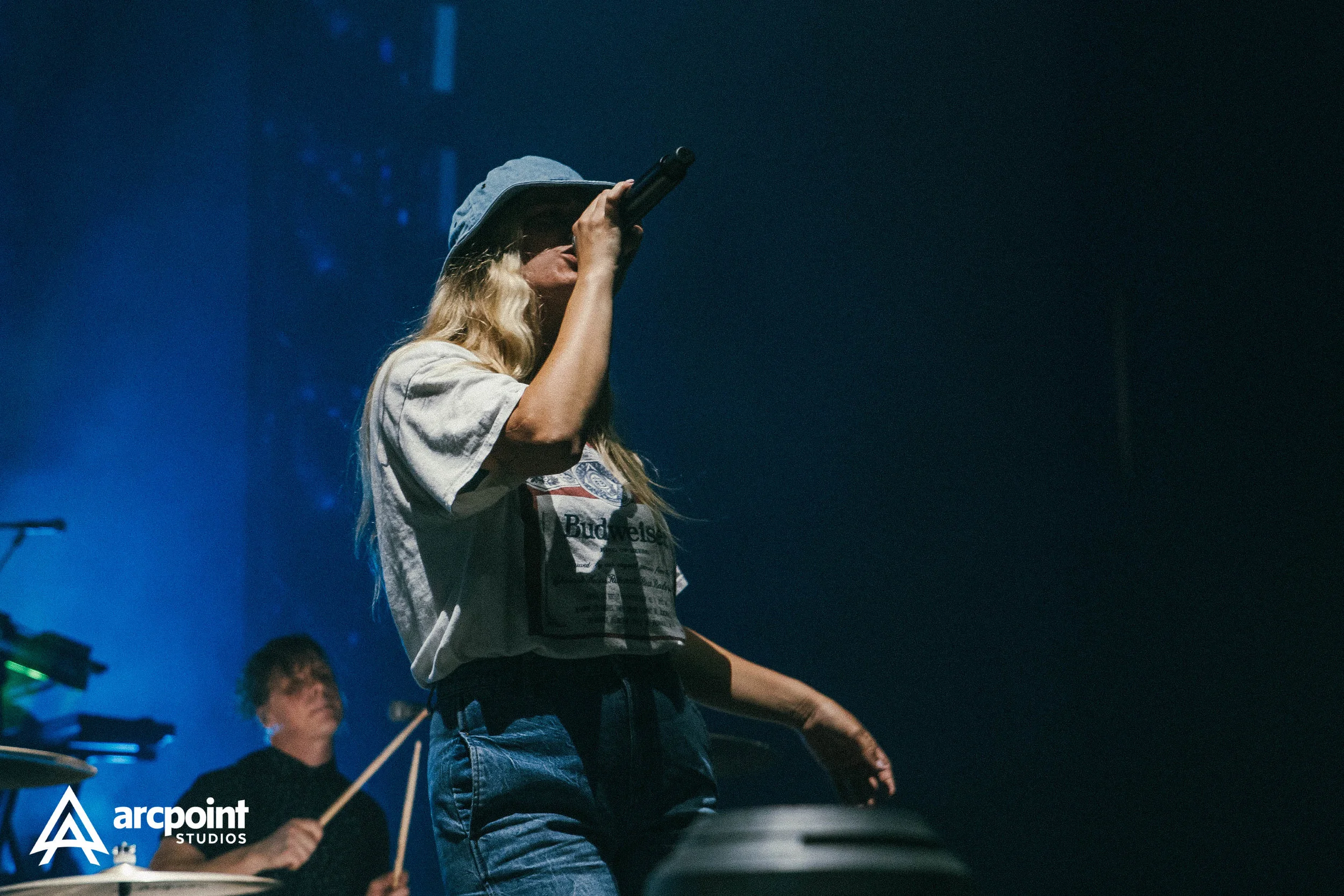 A woman singing into a microphone on stage with a drummer playing in the background, illuminated by blue stage lighting, with the 'arcpoint STUDIOS' logo in the bottom left corner.